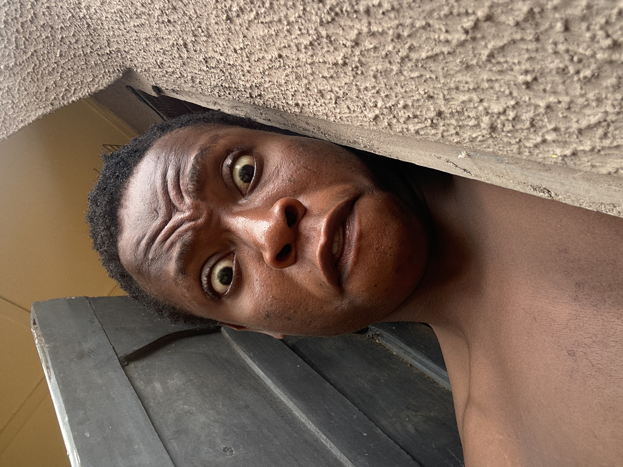 A close-up shot of a person's face from a low angle. The person has dark skin, short, curly hair, and is looking directly at the camera with wide eyes and a furrowed brow, conveying a surprised or questioning expression. The person is positioned near a textured beige surface, possibly a wall, and a dark grey surface that could be a door or another wall. The background is out of focus. The location is Unknown, Unknown.
