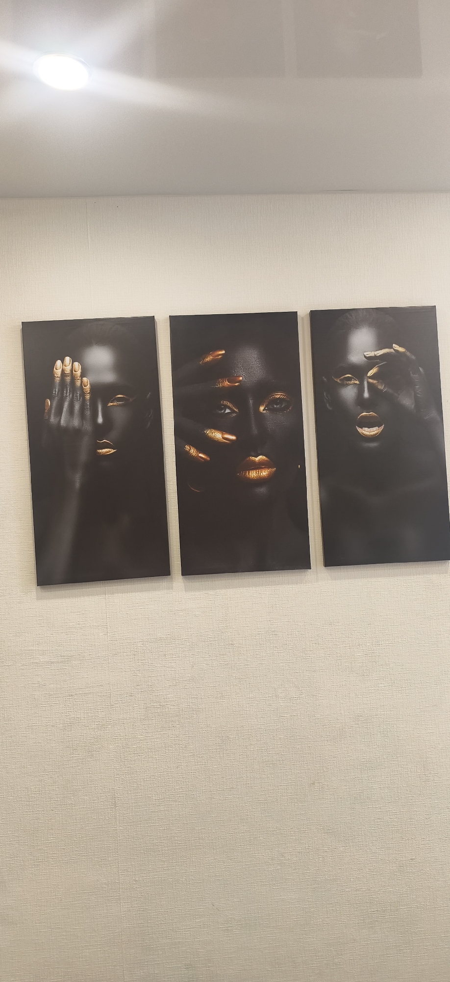 The image shows three framed portraits of women with dark skin and gold accents on their hands and lips, hanging on a beige textured wall. The portraits are positioned horizontally, side-by-side. The location is Unknown, Unknown.