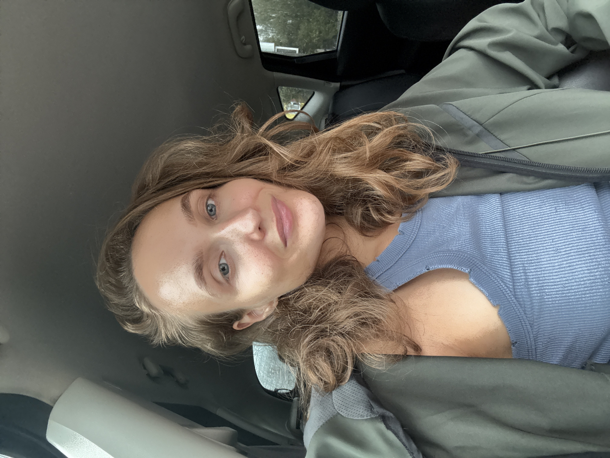 A young woman with light brown hair and blue eyes is sitting in the passenger seat of a car, taking a selfie. She is wearing a blue ribbed tank top and a green jacket. Her hair is wavy and falls around her shoulders. The background shows the interior of the car, including the headrest of the driver's seat and the car window, which reveals a blurry, overcast outdoor scene that appears to be a street in the city of Podshivalovo, Russia.
