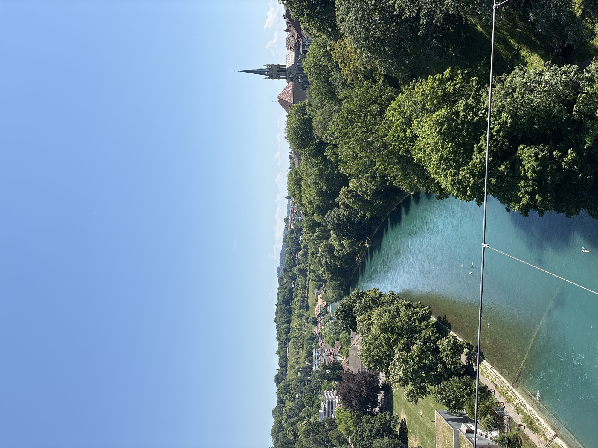 This is an aerial, vertical, wide shot of a scenic river bend with lush green trees lining the banks. The turquoise river water is clear, allowing visibility of the riverbed in shallower areas. A concrete embankment with a paved walkway runs along one side of the river, where a few people are visible strolling. In the river itself, several individuals are swimming or floating, enjoying the water.
On the left bank of the river, a dense forest covers the landscape, with a charming town nestled amongst the trees. Buildings with red-tiled roofs are scattered throughout the hillside. Prominently featured in the distance is a grand church with a tall, pointed spire, characteristic of European architecture. The sky is a clear, bright blue, suggesting a sunny day. The overall scene evokes a sense of tranquility and leisure, with people taking advantage of the beautiful weather and natural surroundings. A thin cable runs diagonally across the upper right portion of the image, hinting at a high vantage point, possibly from a bridge or a cable car. The time of day appears to be midday, given the bright sunlight and lack of long shadows. The location is identified as Bern, Switzerland, which aligns with the architectural style and the presence of a river with this distinctive color and surrounding landscape.