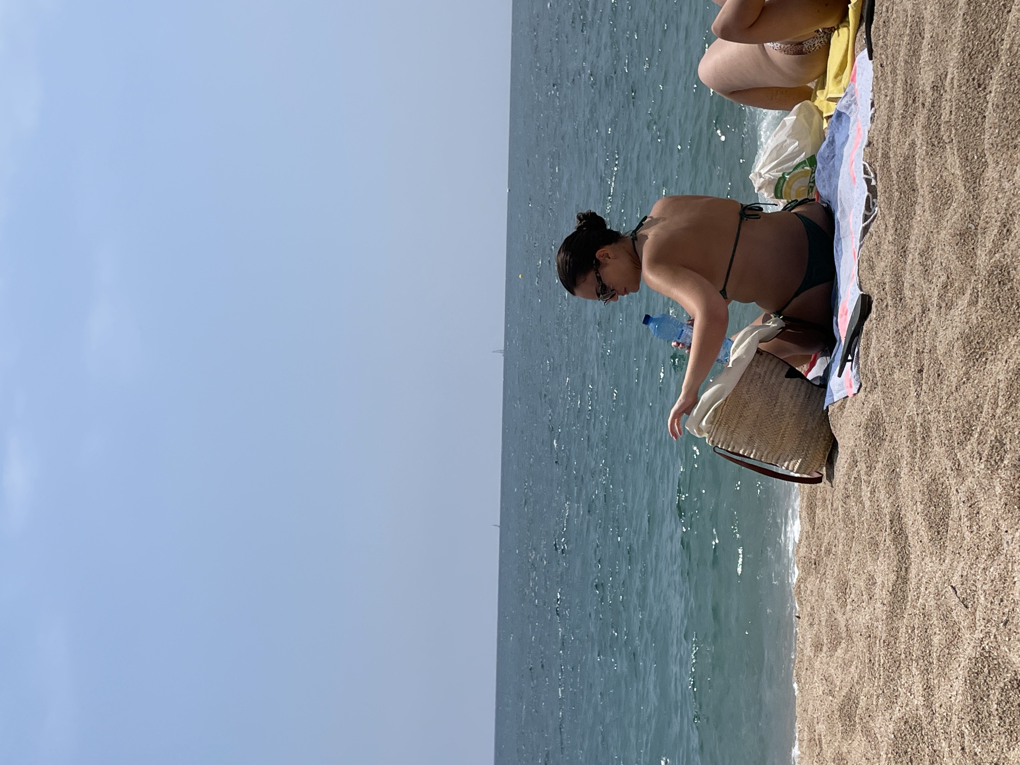 The image depicts a serene beach scene in Canet de Mar, Spain, under a clear blue sky. The foreground is dominated by a sandy beach, with gentle waves lapping at the shore in the background.
A woman, wearing a dark bikini and sunglasses, is seated on a beach towel, facing away from the camera. She is rummaging through a woven straw beach bag. Beside her is a large plastic water bottle and another beach bag, possibly filled with items. To her right, another person, partially visible, is also sunbathing on a yellow towel, wearing a bikini with a leopard print bottom.
The ocean stretches out towards the horizon, with a few distant sailboats visible. The overall atmosphere is one of relaxation and enjoyment of a summer day by the sea.