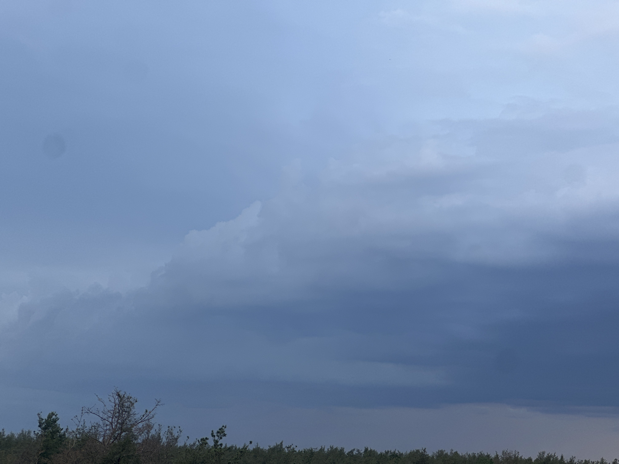 An outdoor, eye-level shot captures a moody sky with heavy, dark clouds dominating the upper portion of the frame. Lighter, wispy clouds are visible in the distance, transitioning to a pale blue sky at the very top. The bottom of the image is lined with a dense, dark green forest, suggesting a natural or rural environment. The lighting is dim, consistent with overcast weather or approaching dusk. The location is Unknown, Unknown.