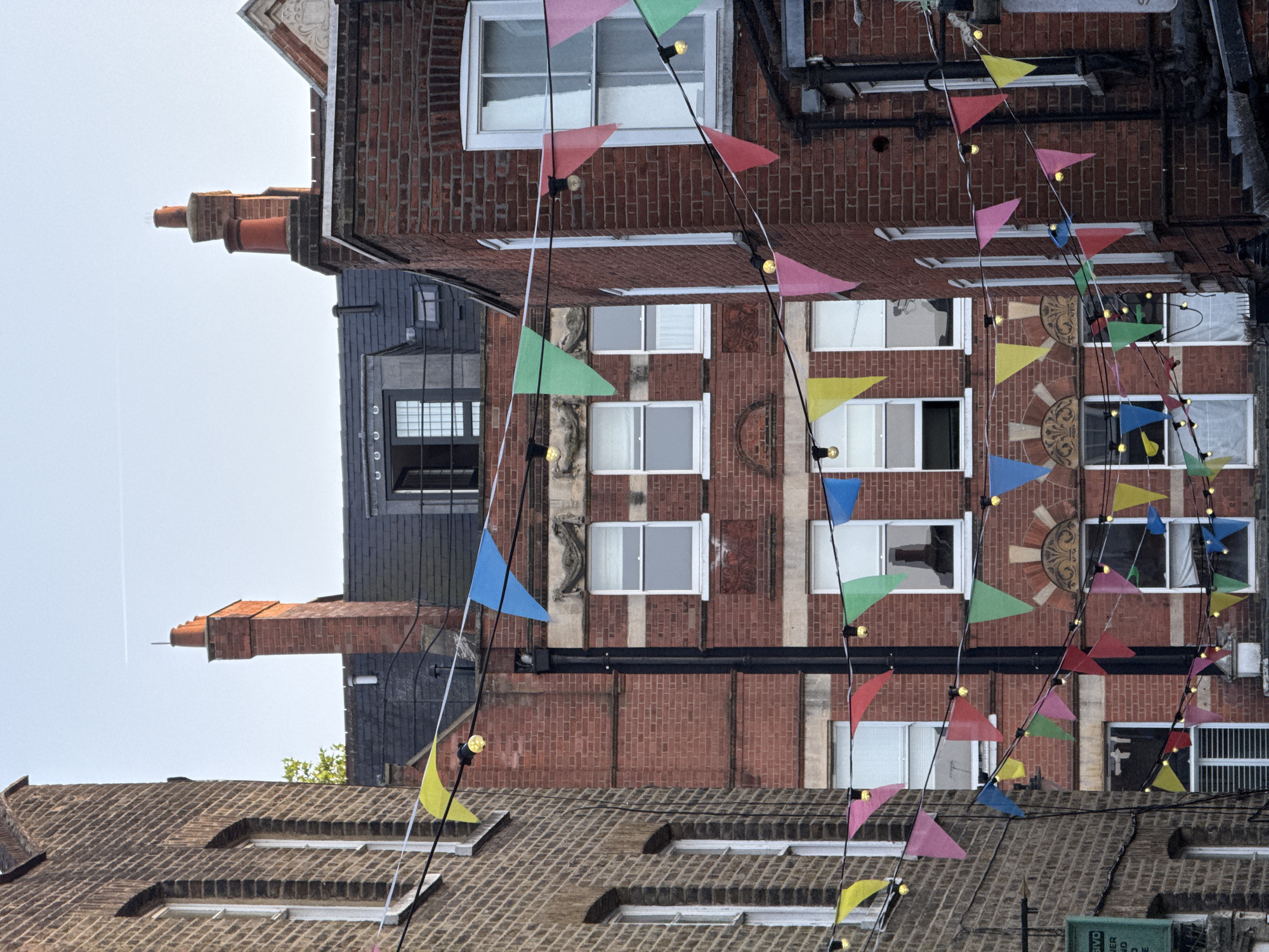 The image shows a street scene in London, United Kingdom. Several brick buildings are visible. Festive bunting with colorful triangular flags and string lights is hung across the street. The sky is light.