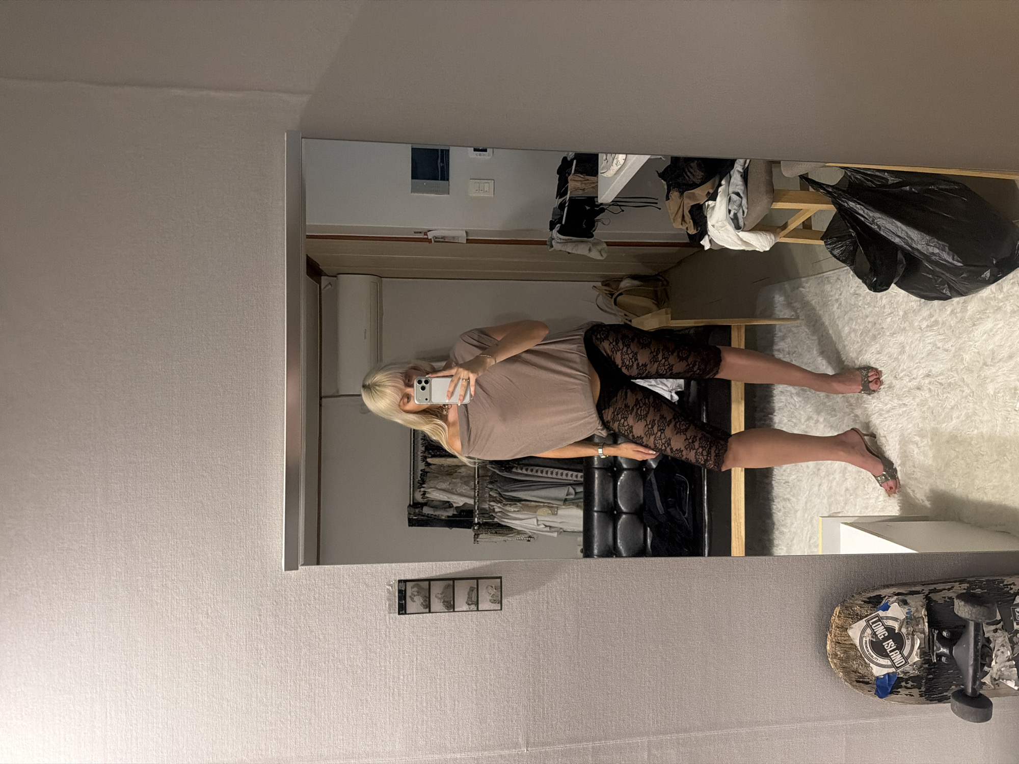A woman is reflected in a mirror in a room. She is holding a smartphone and appears to be taking a selfie. She has blond hair and is wearing a t-shirt and black lace leggings. The room contains a clothes rack with garments, a bench, and a trash bag on the floor. A skateboard is propped against the wall. The location of the image is Seoul, South Korea.
