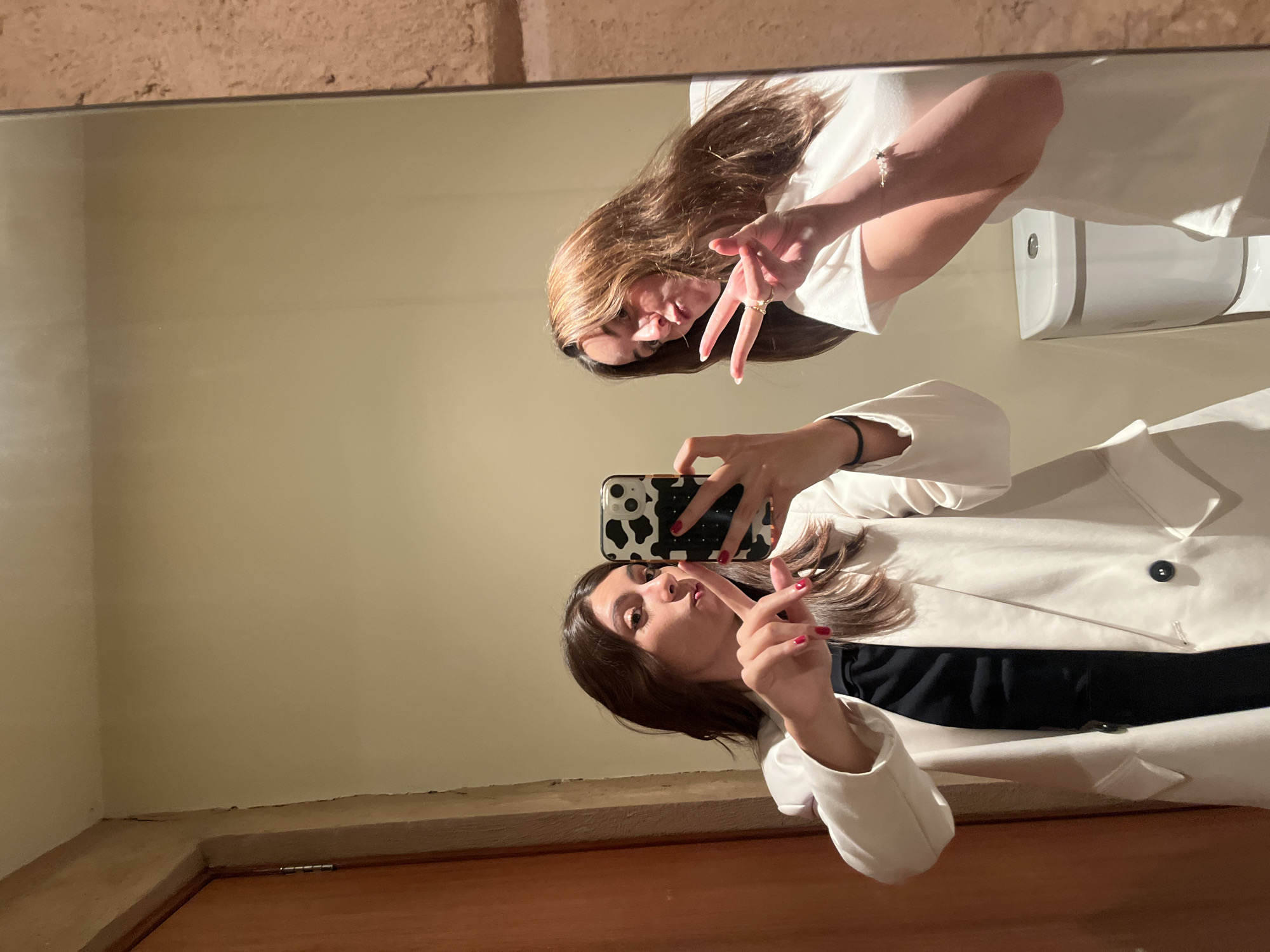 A mirror reflects a woman taking a selfie. The reflection shows a room with a sink and a toilet. The woman is wearing a white blazer over a black top. She is holding a cell phone with a black-and-white cow-print case. She is holding up two fingers in a peace sign. The location is likely a bathroom, possibly in a private residence. The city of San Bernardino Tlaxcalancingo, Mexico, cannot be confirmed from this image.