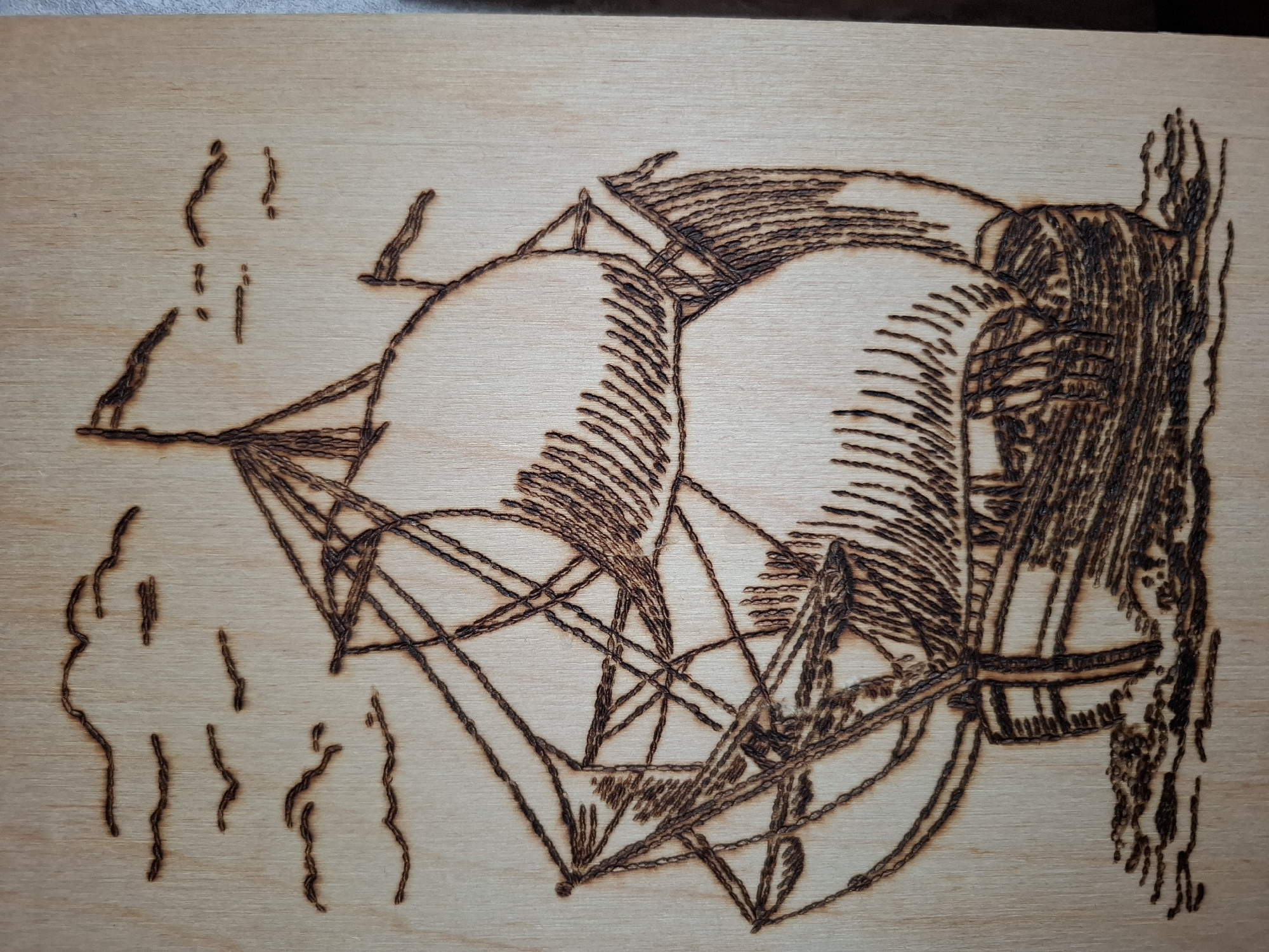 The image depicts a wood-burned drawing on a light-colored wooden surface. The drawing features a galleon ship with prominent sails, rigging, and masts. To the left of the ship are some abstract, vertical lines that may represent smoke or steam. The ship's hull is heavily shaded with dense vertical lines, while the sails are delineated with sparse vertical lines. The rigging consists of numerous intersecting straight lines forming a complex web of triangles. The drawing is rendered in a brown hue from the wood-burning process. There is no information related to location in the city of Амзя, Russia.