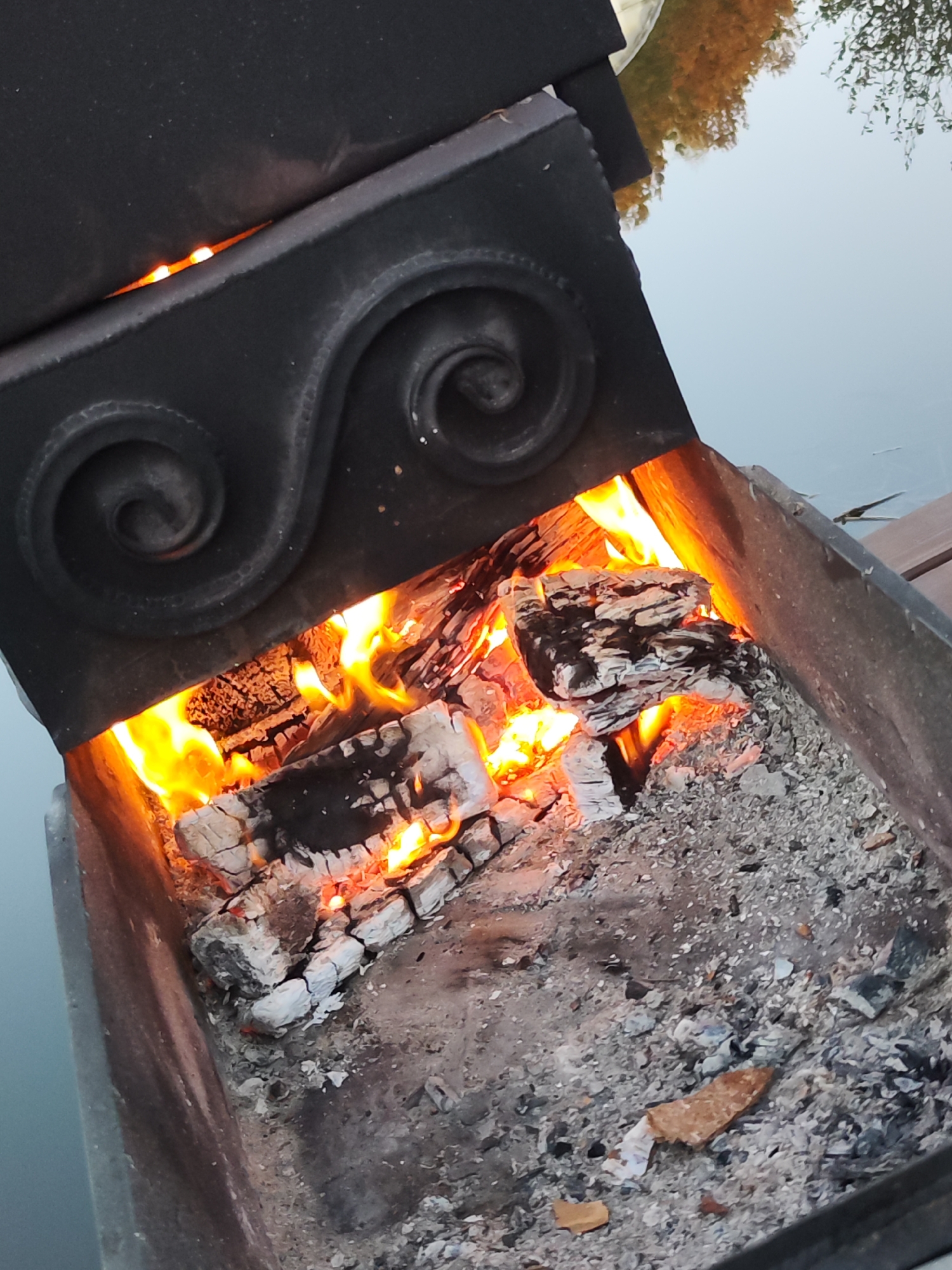 A close-up, angled shot depicts a black metal fire pit containing burning wood. Embers and ash line the bottom. The upper portion of the fire pit has decorative scrollwork. Reflections of trees and what appears to be water are visible in the upper-right corner. The specific location is likely Chaikovsky, Russia, based on the prompt's instructions.