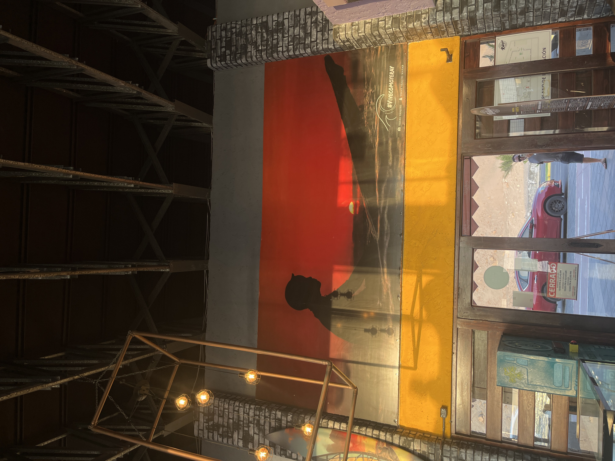 Here's a detailed description of the image:
**General Description:** The image shows an indoor scene with architectural elements and decorative features. The perspective is slightly angled, giving a view upwards and outwards towards a doorway.
**Architectural Elements:**
* **Ceiling:** A complex structure of metal beams forms a grid-like pattern on the ceiling.
* **Walls:** Brickwork details are visible along the top of the walls, where they meet the ceiling. A large, colorful poster or mural is displayed on the wall.
* **Doorway:** A wooden-framed doorway leads to an outdoor area. Signage is visible on the doorway, along with a "CERRADO" sign.
**Decorative Features:**
* **Lighting:** A copper-colored metal framework with exposed light bulbs hangs in the foreground.
* **Mural/Poster:** A large artwork with red, orange, and yellow hues features a silhouette of a person. The text "#VIVECOMOYERAY" is visible on it.
**Outdoor Scene Visible Through Doorway:**
* **Cars:** A red car is partially visible outside.
* **People:** A person is seen standing outside.
* **General Environment:** A glimpse of the street and buildings in the background is seen.
**Contextual Information:**
* **Text on Poster:** The poster includes the hashtag "#VIVECOMOYERAY" and names "YERKO VÁSQUEZ" and "YERAY VÁSQUEZ MAHAN".
* **Signage:** The signage on the doorway includes the name "Rūky's" and the words "PLANO DEL".
**Inference (Based on text and visual clues):** This is likely the interior of a business (perhaps a restaurant or bar) called "Rūky's". The artwork appears to be related to a promotion or campaign, possibly associated with the names Yerko Vásquez and Yeray Vásquez Mahan. The "CERRADO" sign suggests the establishment is currently closed.
**Location:** While the image doesn't explicitly state the city, based on the OCR text and the architectural style, it may be located in Arica, Chile.