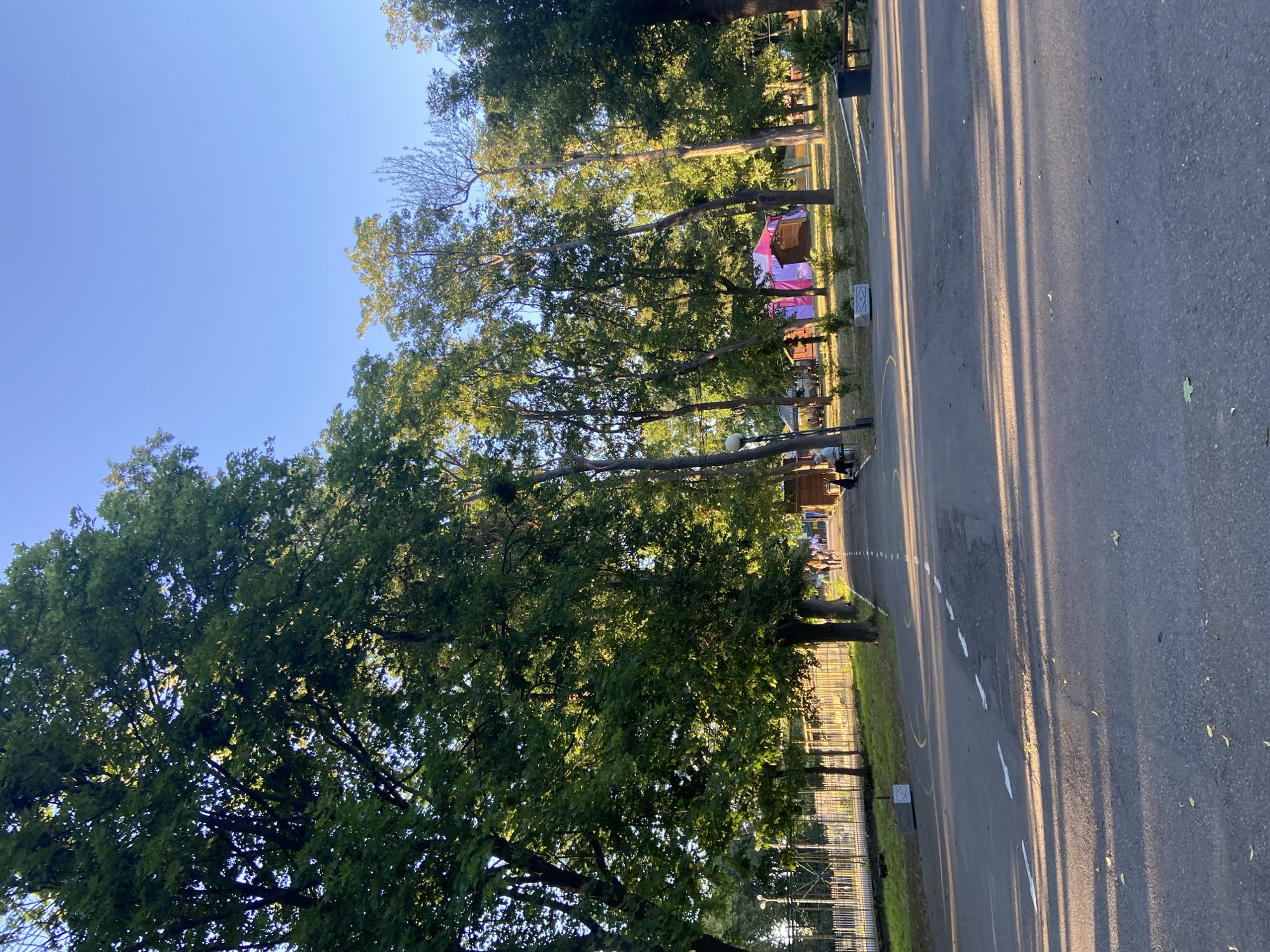 The image depicts an outdoor scene on a sunny day, likely late afternoon due to the long shadows cast across the asphalt path. The path runs along the right side of the frame, with a grassy area and trees to its left. In the distance, beyond the trees, there appear to be some structures, including what looks like a tent or booth with pink and white fabric. Several people are visible near these structures, engaged in some activity, though their specific actions are not clear. A fence runs along the edge of the grassy area. The overall setting suggests a park or recreational area. The sky is a clear blue. The image is oriented vertically, with the sky at the top and the ground at the bottom. There is no discernible text in the image.