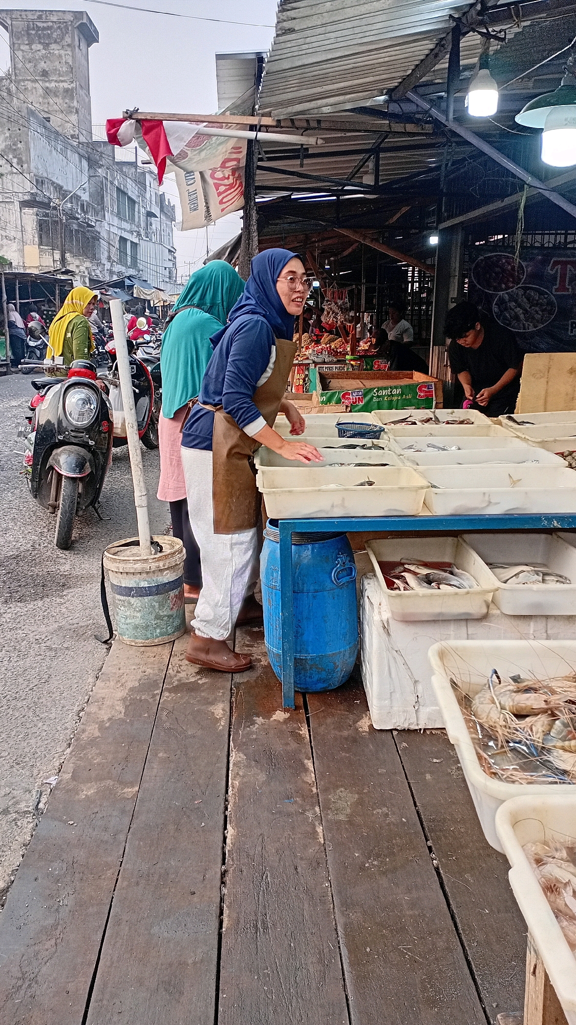 A street market scene is depicted in Jambi City, Indonesia, on a cloudy day. In the foreground, a woman wearing a blue hijab and a brown apron is sorting through fish displayed in white plastic trays on a blue shelf. She is barefoot and wears light-colored pants. The wooden planks of the market stall are wet, suggesting recent cleaning or rain. To her left, a blue plastic drum sits on the planks. Behind her, other market-goers and motorcycles are visible, along with buildings in the background that appear aged. To the far right, another person is visible behind a stall, possibly engaged in a transaction. The market is characterized by its rustic wooden structures and an abundance of fresh produce, primarily fish, laid out for sale.