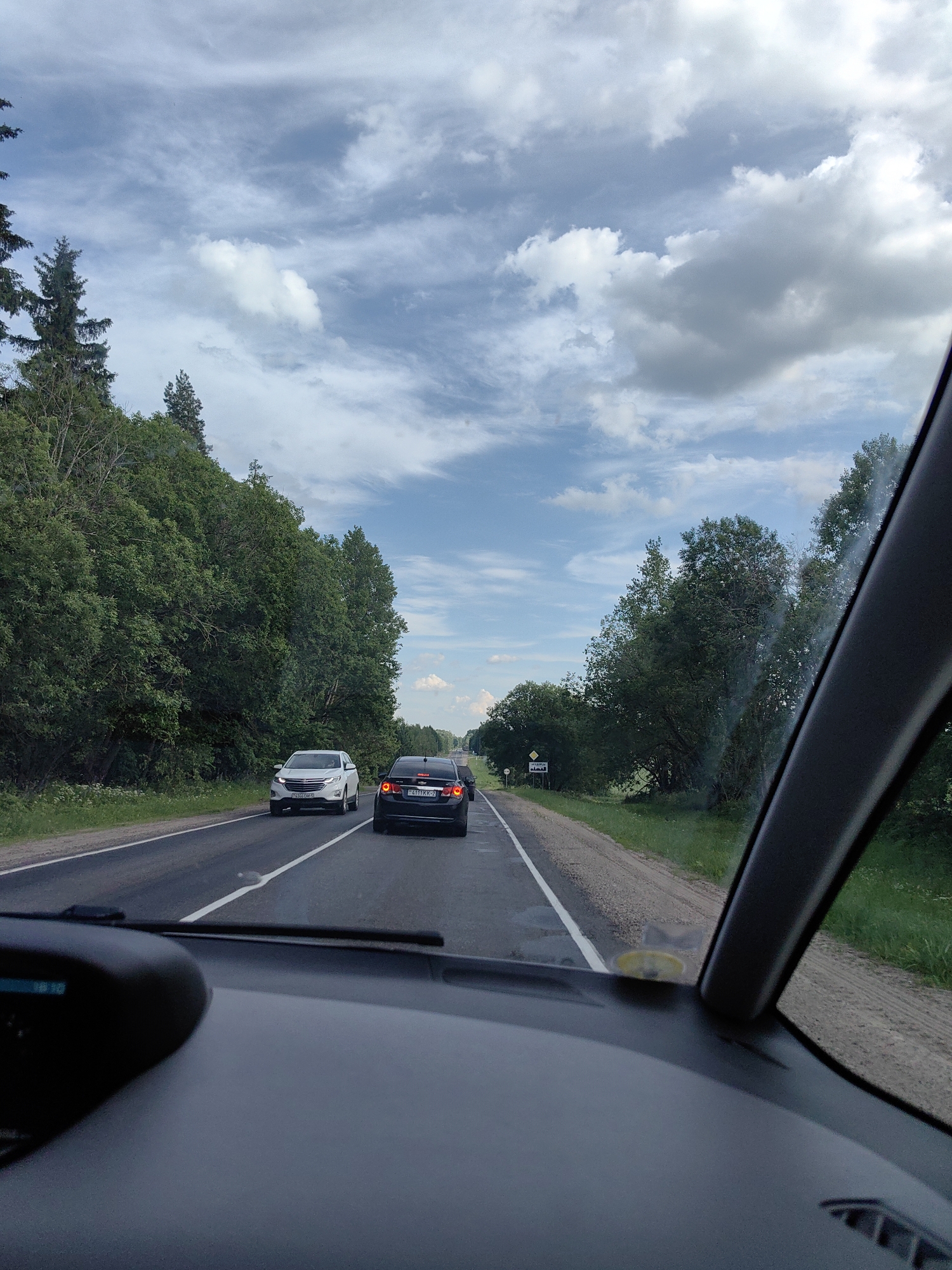 This is a vertical, eye-level, daytime, full-shot photograph taken from the interior of a vehicle traveling on a paved road. The road runs through a rural landscape with dense green trees lining both sides. The sky is partly cloudy with scattered white and grey clouds against a light blue backdrop.
In the foreground, the dashboard of the vehicle is visible, along with a portion of the windshield frame on the right. On the road ahead, several cars are visible. A white SUV is in the left lane, and a dark-colored sedan is in the right lane, followed by another dark-colored vehicle. The cars are traveling in the same direction as the photographer.
The scenery suggests a journey through a forested or heavily treed area. There is a yellow diamond-shaped road sign with black symbols on the right side of the road. The environment appears to be the territory of Ilyanskiy selskiy Sovet, Belarus, given the presence of a sign with Cyrillic text that reads "Ільянскі сельскі Савет" (Ilyanskiy selskiy Sovet) and "Вязынка" (Viazynka), indicating the geographical location.