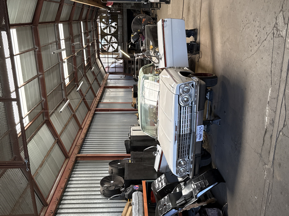 This image depicts the interior of an industrial workshop or garage in Los Angeles, California, United States of America. The primary subject is a light-colored (likely white or light grey) 1962 Chevrolet Impala convertible with its top down, positioned centrally in the foreground. The vehicle is elevated on a single jack stand visible beneath its front bumper, with its front wheels off the ground. A California license plate, "9WWT925," is affixed to the front bumper. The car features chrome trim, a prominent grille, and red wheels with whitewall tires.
A single individual is observed inside the driver's side of the Impala, leaning into the interior. They are wearing a dark welding helmet or face shield, dark clothing, and gloves, actively engaged in work within the vehicle. The driver's side door is open, revealing the person's feet and lower legs on the concrete floor, adjacent to a dark equipment box.
The environment is characterized by a high, arched corrugated metal roof supported by red-brown painted steel trusses. Translucent skylight panels and several suspended fluorescent tube lights provide illumination. The walls are constructed from corrugated metal panels. On the left side of the image, stacks of large industrial fans are visible against the wall, along with various containers including a black fuel can with an orange "NP" logo. Several dark, reflective automotive body panels or parts are propped against a shelf in the left foreground. The concrete floor shows signs of wear and cracks. In the background on the right, additional large industrial fans and machinery are discernible, suggesting an active workspace or fabrication area, with a raised platform or mezzanine visible further back.