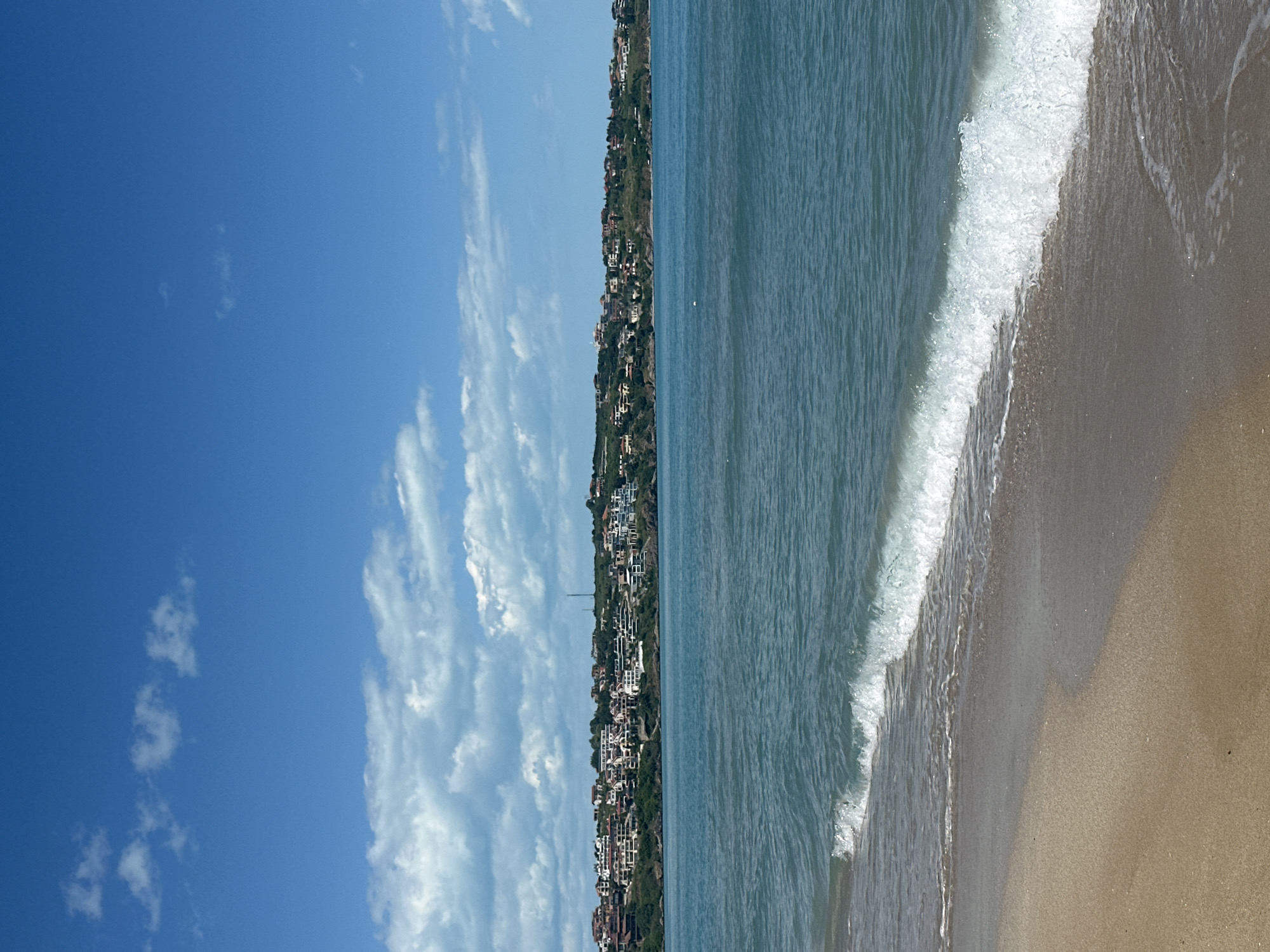 The image depicts a coastal scene in Sozopol, Bulgaria. The scene is comprised of a blue sky with scattered cumulus clouds, a coastline with buildings on a cliff, the Black Sea, and a sandy beach with waves. The coastline has many buildings and vegetation on the cliffs above the sea. The sea is a dark blue-grey in color, with waves crashing on the shore. The beach is a light tan color.