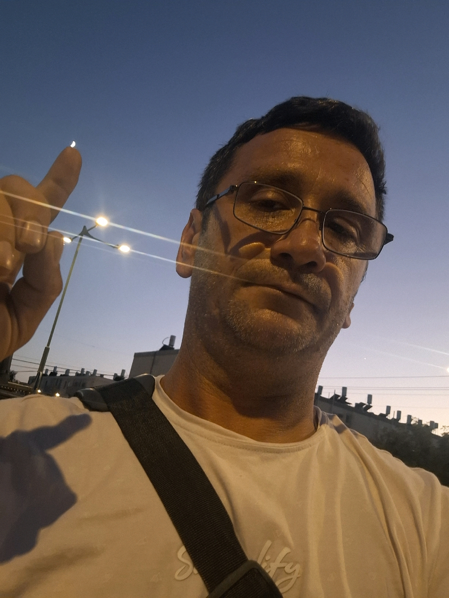 A man wearing glasses and a white t-shirt is taking a selfie outdoors at dusk in Yerocham, Israel. He is pointing his left index finger towards a sliver of the moon in the clear blue sky. In the background, streetlights illuminate the scene, with buildings visible in the distance. The man's expression is neutral as he looks directly at the camera.