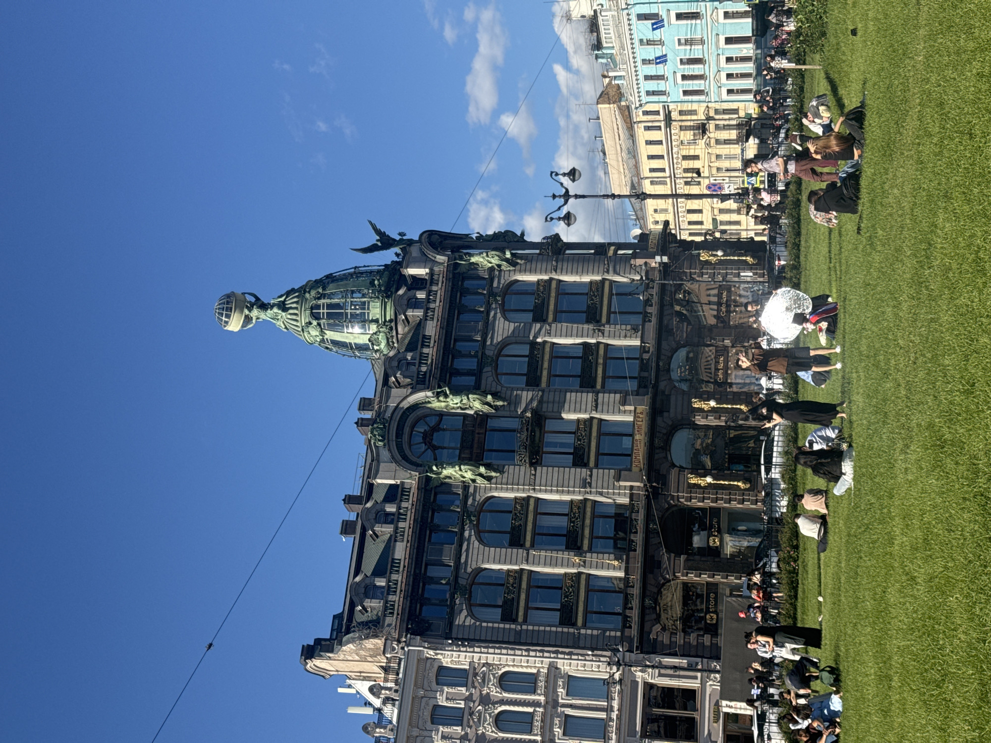 The image depicts the Singer House, also known as the Dom Knigi (House of Books), a prominent Art Nouveau building located on Nevsky Prospekt in Saint Petersburg, Russia. The building is characterized by its distinctive glass dome topped with a globe supported by an eagle sculpture. In the foreground, a vibrant green lawn is dotted with numerous people of various ages, many of whom are seated or reclining, enjoying the sunny day. Some individuals are engaged in conversations, while others appear to be relaxing or posing for photographs. A photographer is visible on the left side of the lawn, utilizing a large circular diffuser to illuminate their subjects. The background showcases the Singer House and other colorful buildings lining the street, along with streetlights and a clear blue sky with scattered clouds. The scene exudes a sense of leisure and urban life on a pleasant day in Saint Petersburg.