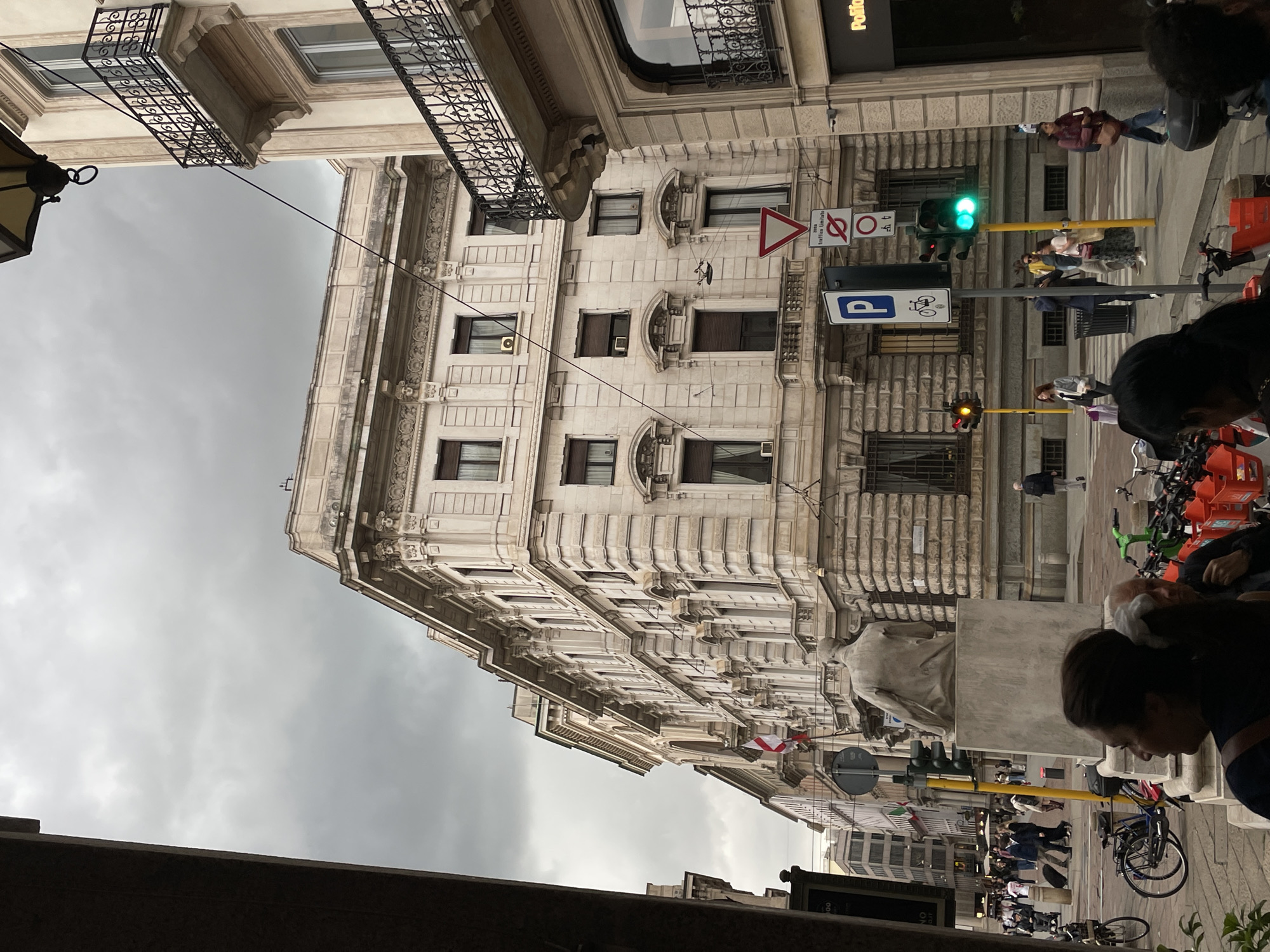 The image depicts a street scene in Milan, Italy, notable for its architecture and urban activity. A multi-story building with ornate façade details stands prominently, its windows and architectural elements visible against a cloudy sky. Pedestrians are present on the sidewalk, interacting with the environment. A statue stands near a traffic light. Bicycles are parked along the street, and traffic lights are in operation. Signage is present, indicating local regulations. The scene is typical of a European urban environment, with a mix of historic buildings and modern urban infrastructure.
