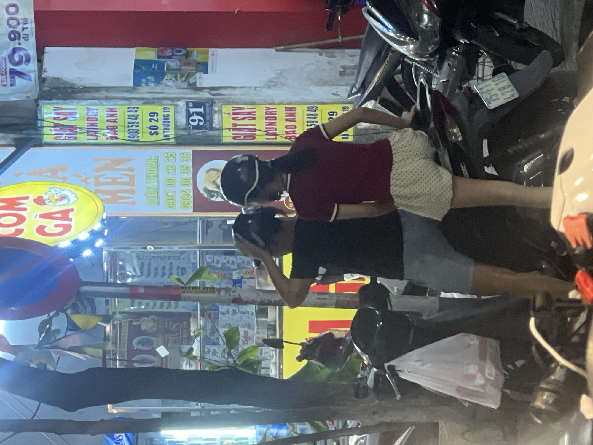 The image depicts a street scene at night in Đà Nẵng, Vietnam, likely near a commercial area. In the foreground, two individuals are seen on or near motorcycles. One person, wearing a black t-shirt and blue shorts, is standing and appears to be adjusting their helmet. The other person, wearing a maroon t-shirt and white polka dot shorts, is seated on a motorcycle. Both individuals are wearing helmets, suggesting adherence to local traffic regulations.
Several signs are visible in the background. A prominent yellow sign with red text reads "GIẶT SAY LAUNDRY BẢO ANH 0903 59 48 49 SỐ 29 AN THƯỜNG 19". Another sign, also yellow, features Vietnamese text and a graphic of a chicken, possibly indicating a food establishment. A blue and red traffic sign, likely a prohibition symbol, is also present. The environment appears to be urban, with streetlights illuminating the scene and other motorcycles parked nearby. A white plastic bag containing items is hanging from the handlebars of one of the motorcycles. The overall atmosphere is that of a bustling city street at night.