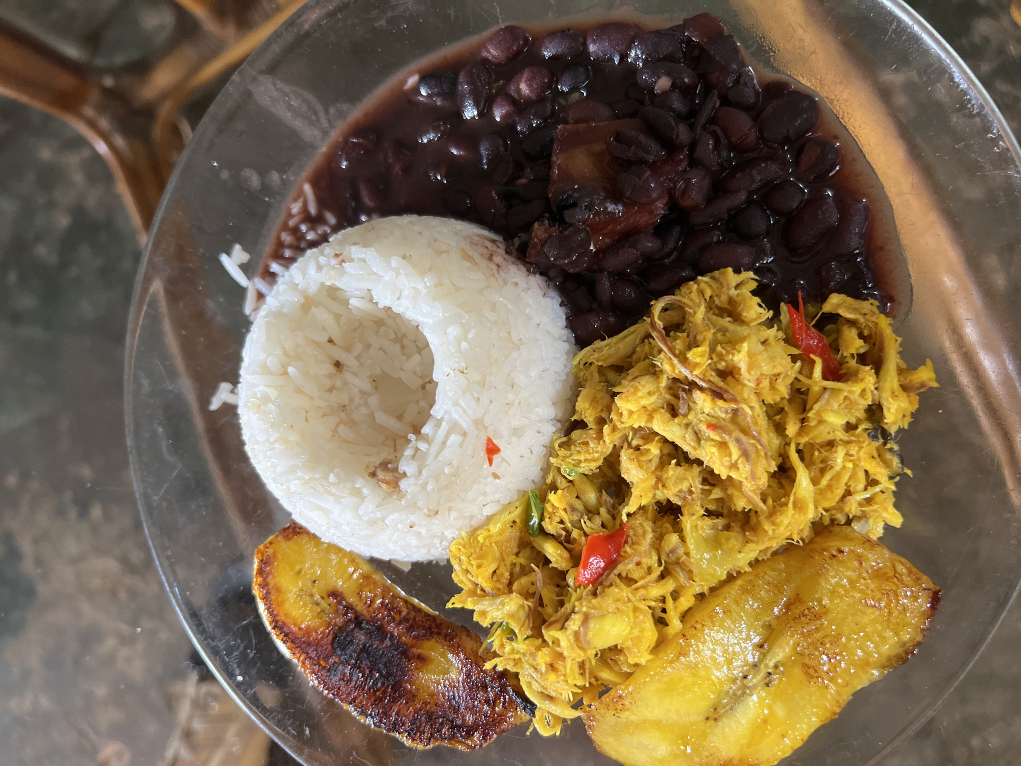 A close-up, overhead shot shows a circular glass plate filled with food. The plate is situated on a dark, textured surface, possibly a wooden table. The food consists of several distinct components arranged in sections.
At the top center is a mound of fluffy white rice, with a small indentation in the center. To the right of the rice are dark red beans, some appearing whole and others mashed, in a rich dark sauce. There are also a few pieces of what looks like pork or meat in the bean mixture.
To the left of the rice, there is a portion of shredded yellow meat, seasoned with spices and possibly containing red chili peppers. Adjacent to this shredded meat, at the bottom left of the plate, are two slices of fried plantain, one at the bottom and one to the left of the shredded meat. The plantains are golden brown and appear slightly caramelized.
The lighting suggests an indoor setting, likely a restaurant or home, with a blurred background that hints at wooden furniture, possibly a chair or table legs, in a dark brown hue. The image composition emphasizes the textures and colors of the food. The scene depicts a prepared meal, ready to be eaten, in Juan Griego, Venezuela.
