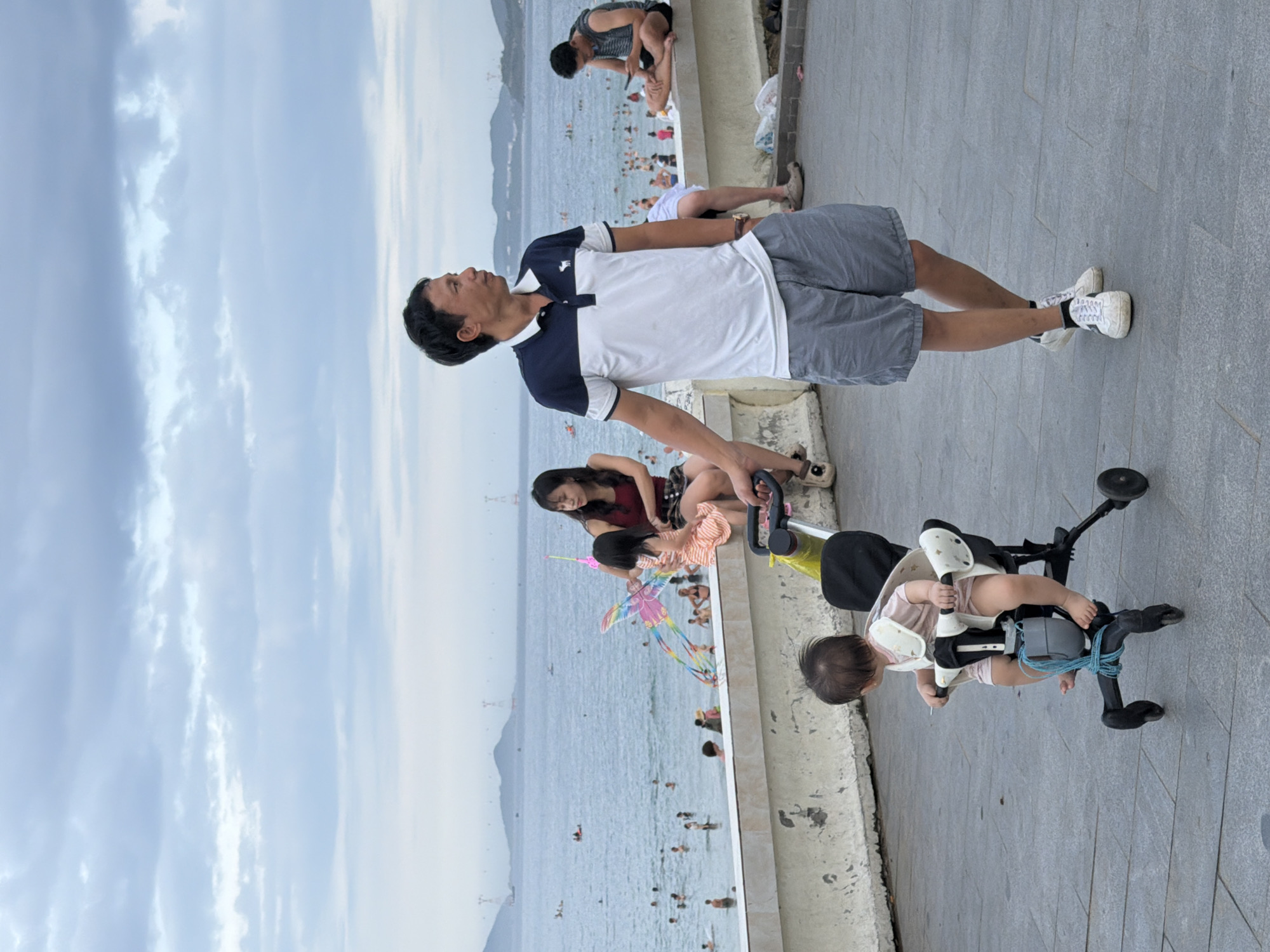 In Nha Trang, Vietnam, a man in a blue and white polo shirt and grey shorts walks along a paved promenade next to a beach. He is looking towards the sea, which is filled with swimmers and a few boats. In the foreground, a baby sits in a stroller, facing away from the camera, secured by a blue rope. To the left of the man, a woman in a red top and patterned skirt sits on a low wall, holding a young child who is reaching for a colorful kite. Another woman is partially visible on the wall to the far right, looking at a phone. The sky is overcast, with faint mountains visible in the distance.