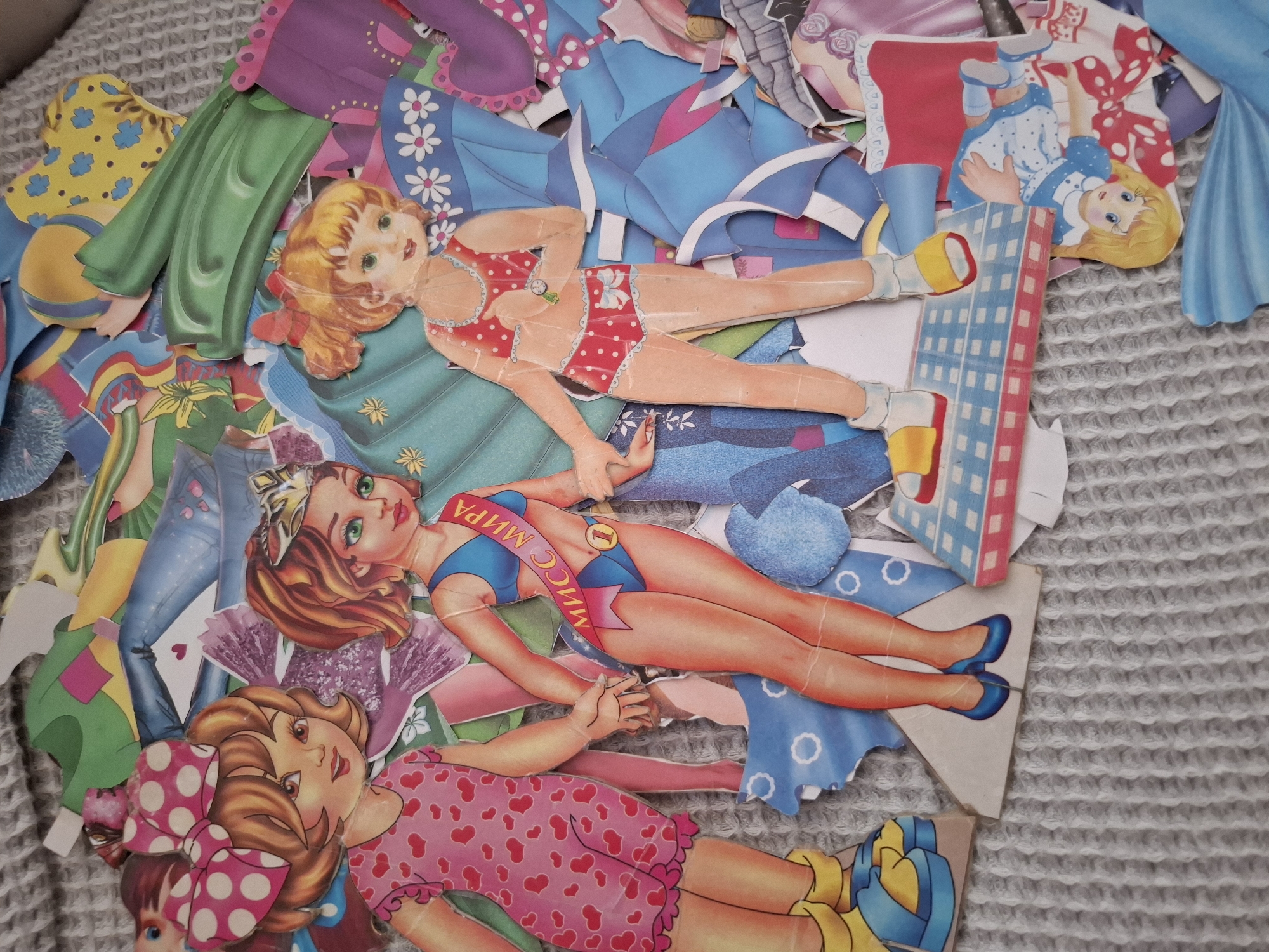 The image shows a collection of vintage paper dolls and their clothing scattered on a gray textured surface. The dolls are drawn in a retro style, with bright colors and detailed outfits. One doll wears a red and white polka dot swimsuit, another a blue swimsuit with a "Miss Mira" sash written in Russian. The clothing items include dresses, skirts, pants, and accessories. A building is also present. The location, Amzya, Russia, is obtained from the sash Мисс Мира (Miss Mira, meaning Miss World).