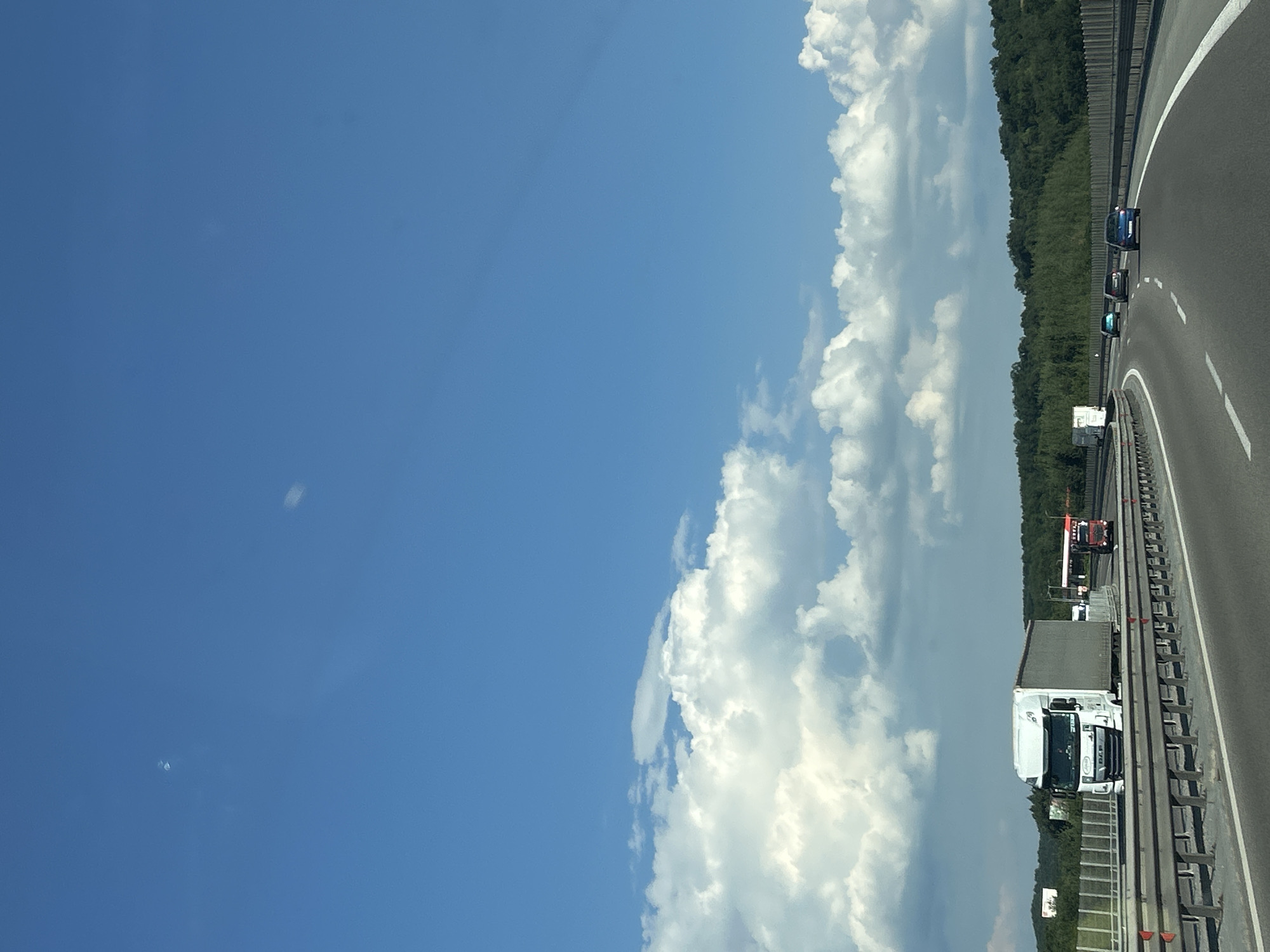 This image shows a highway scene with a clear blue sky and white clouds. The highway appears to be a multi-lane road with a guardrail separating it from the adjacent landscape, which is filled with lush green trees. Several vehicles are visible on the road, including a white truck with a trailer, a red and black truck, and a few cars. The vehicles are traveling in the same direction. The perspective suggests the photo was taken from another moving vehicle. The weather appears to be sunny and pleasant, suggesting it's daytime. There are no visible people, but the presence of vehicles indicates human activity. The location is identified as Baliovtsi, Bulgaria, which would imply a European setting. There is some text visible on the white truck's trailer, but it is too blurry to read. The overall impression is a typical highway scene on a clear day.