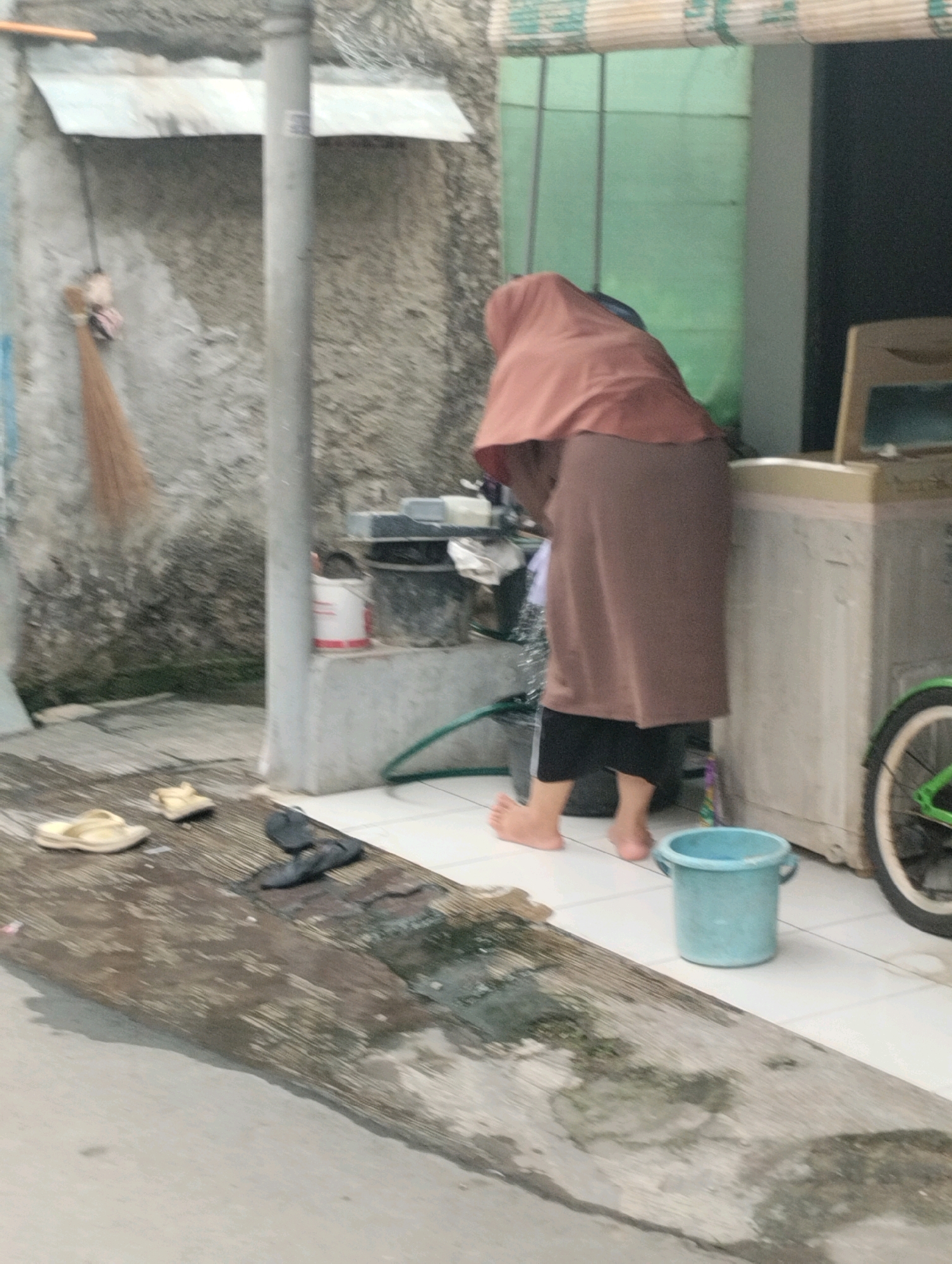 In Bekasi, Indonesia, an outdoor scene unfolds with a person wearing a brown headscarf and a long brown skirt, with black trousers underneath. The individual is positioned near a cream-colored appliance, possibly a washing machine, and appears to be engaged in an activity by a sink. The ground surface is tiled, with a wooden platform situated in the foreground. This platform is wet, with some debris on it, and is adjacent to a concrete path. A light blue bucket is visible on the tiled ground. In the background, a concrete wall with a corrugated metal awning is present, and a broom hangs on the wall. A bicycle with a green rim is partially visible to the right of the scene.