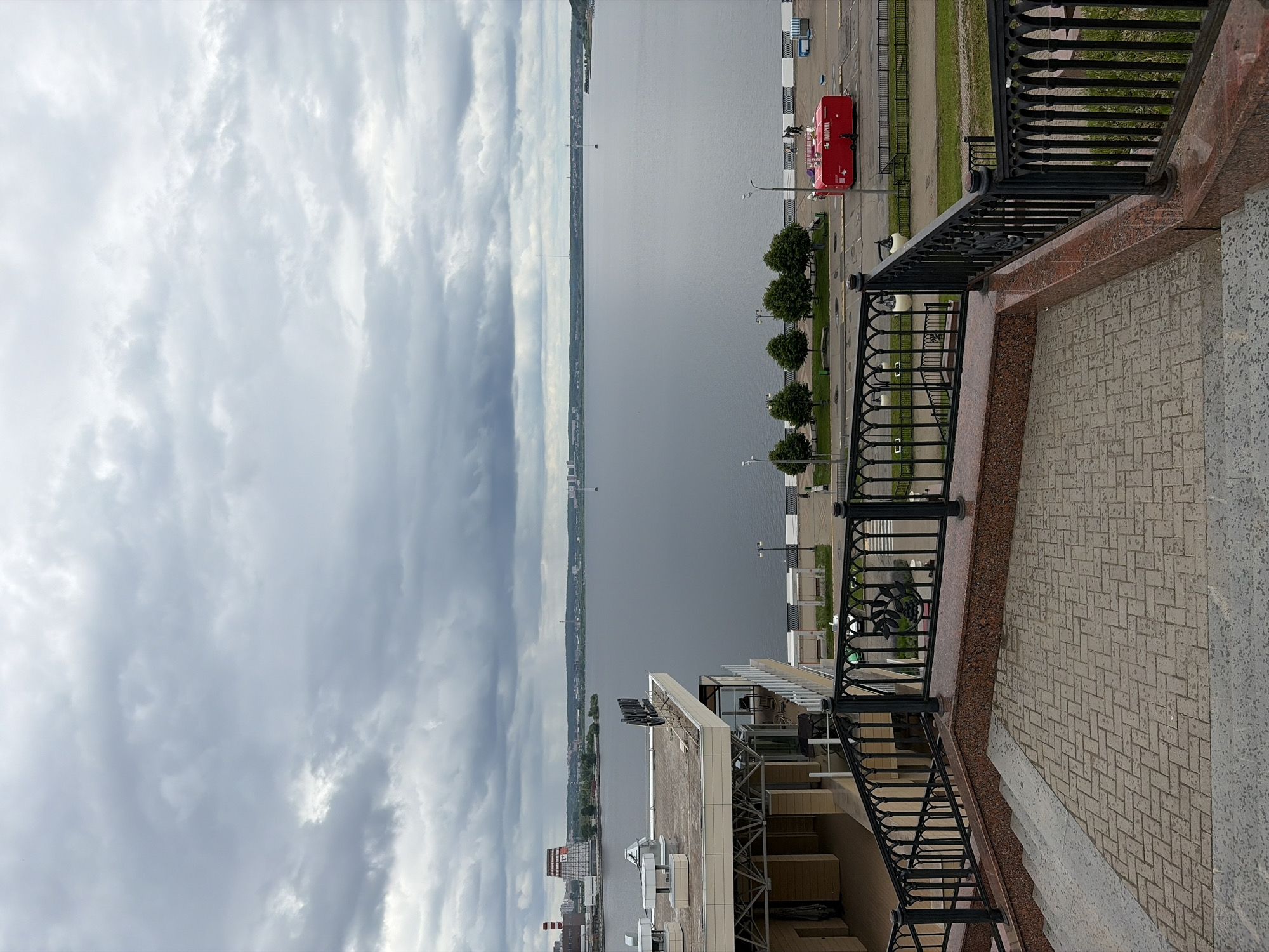 The image presents an elevated view overlooking a wide body of water, likely a river or large lake, under a heavily overcast sky. In the immediate foreground, a black metal railing with ornate details runs along a pathway or balcony, which features light-colored patterned paving tiles and a reddish-brown marble-like balustrade.
Beyond the railing, a paved promenade extends along the near bank of the water. It is sparsely lined with small, evenly spaced green trees and streetlights. A prominent bright red food truck or trailer, displaying the Cyrillic text "ПЛОВМАН" (Plovman), is parked on the promenade, with a few small, indistinct figures visible nearby.
The vast expanse of the water occupies the midground, appearing calm and reflecting the grey, cloudy sky. On the opposite bank, a distant shoreline with various buildings and structures suggests urban development stretching into the horizon. To the lower left, part of a building with a flat roof and modern architectural elements is visible, indicating the structure from which the photo was taken.
The sky is filled with thick, grey clouds, suggesting an overcast day, possibly late morning or afternoon, without direct sunlight. The scene is calm and depicts an urban waterfront environment. The location is Izhevsk, Russia.