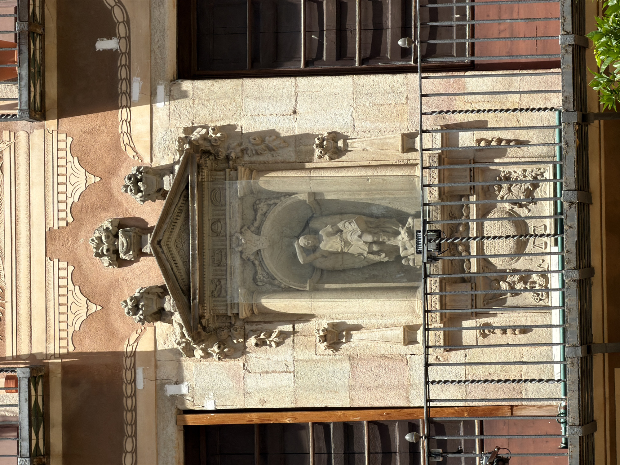 The image shows a close-up view of an ornate architectural detail on the facade of a building, likely in Barcelona, Spain, given the style of the architecture and balcony railing. In the center is a stone niche containing a sculpture of a male figure with wings, possibly an angel or a saint, holding a sword above his head and standing over a fallen figure. The niche is framed by a decorative arch and pilasters. Above the niche, there are carved decorative elements including what appear to be lion heads and floral motifs. The niche is protected by a transparent sheet of glass. A wrought-iron balcony railing with decorative twists is in the foreground, partially obscuring the lower part of the architectural feature. The building's facade is made of light-colored stone and decorated with patterned plasterwork in shades of orange and brown. To the left and right of the central feature are tall, dark wooden window frames. The overall impression is one of historical architecture with detailed craftsmanship.