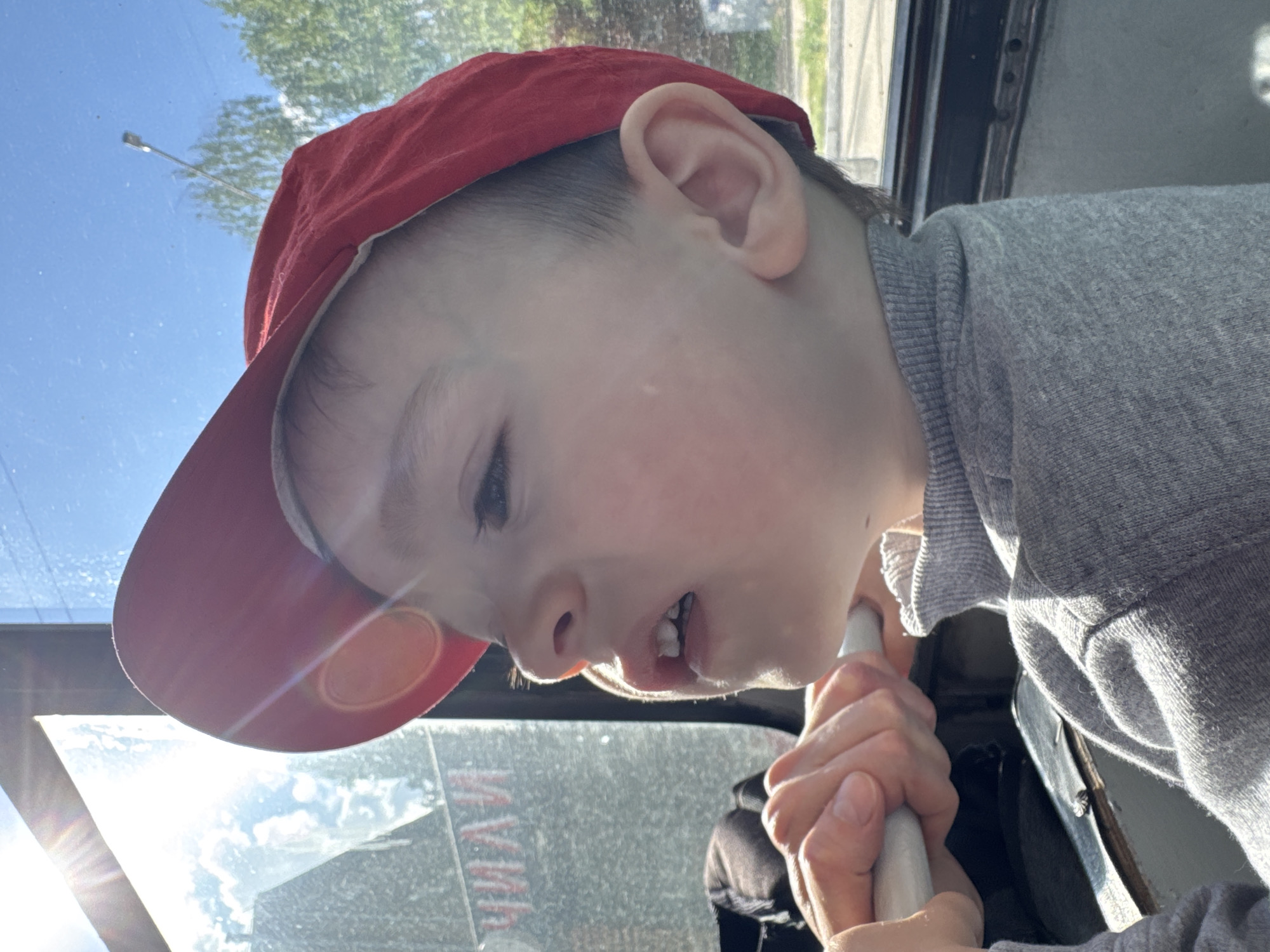 A young child, wearing a red baseball cap and a gray long-sleeved shirt, is seated inside a vehicle, likely a bus or truck, based on the visible interior paneling and window frame. The child is holding onto a white metal bar, possibly a handrail. The background visible through the window shows a partially obscured urban scene with greenery, a street light, and possibly buildings. The city is Izhevsk, Russia. Sunlight is reflecting off the windshield.