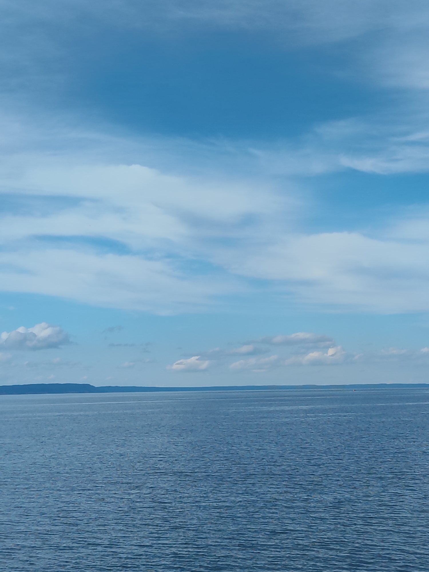 A wide shot captures a vast expanse of calm, blue water stretching towards a distant, dark blue landmass under a bright sky. The water's surface is subtly rippled, reflecting the light. Scattered white clouds drift across the light blue sky, some wispy and thin, others more substantial. The horizon line is clearly defined between the water and the land. The scene appears serene and undisturbed. There are no people or man-made structures immediately visible, except for a tiny, distant boat on the right side of the image. The location is identified as Unknown, Unknown.