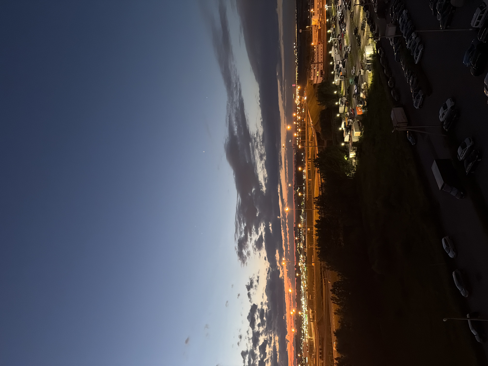 The image shows a city scene at dusk. The sky is a gradient of deep blue to orange, with scattered clouds. In the distance, there are city lights and buildings, and a large parking lot with many cars. A highway with traffic lights is visible in the middle ground. In the foreground, there is a dark area with trees and a few parked cars. The overall impression is one of an urban landscape transitioning from day to night. The specific location is Unknown, Unknown.