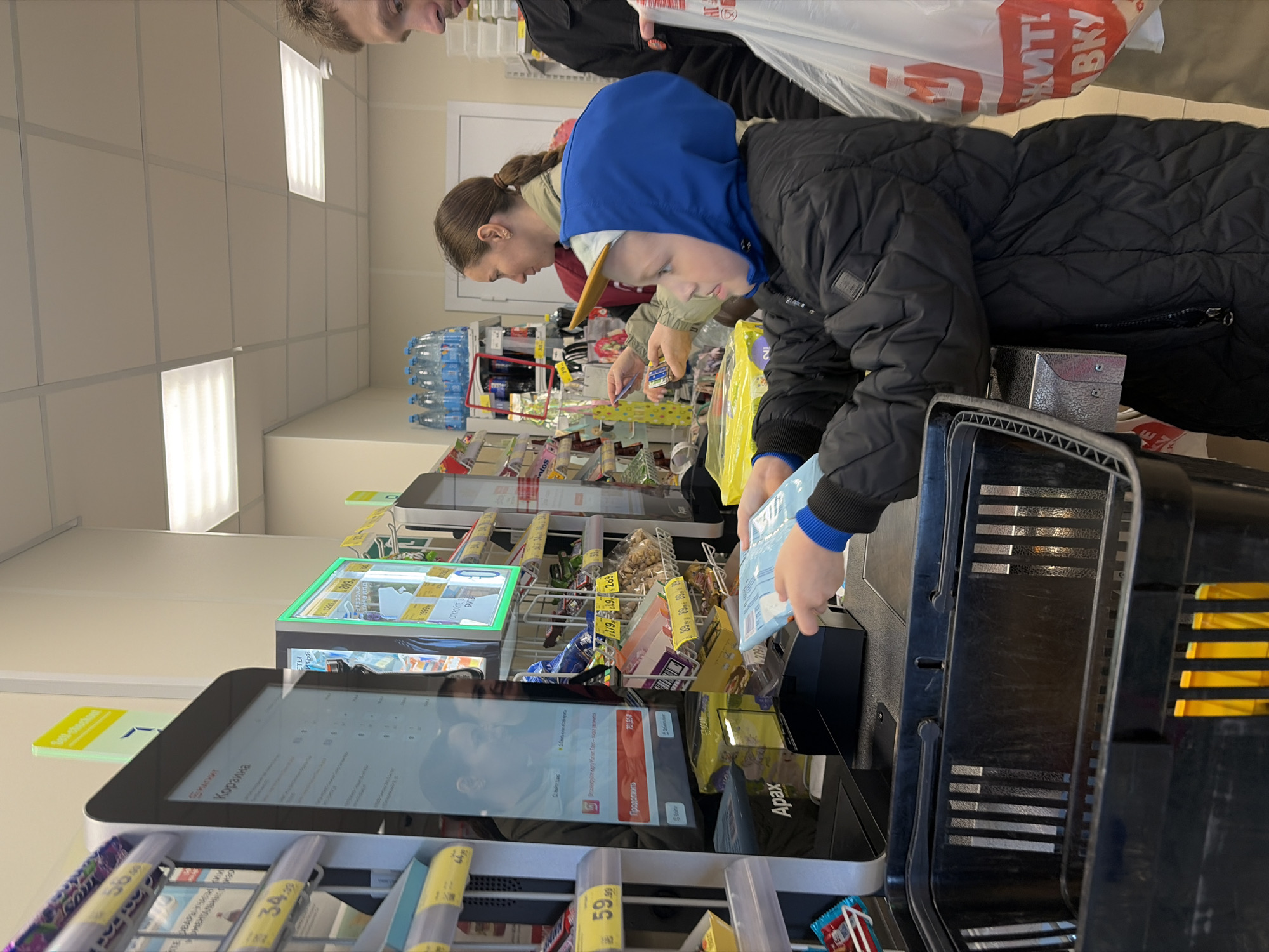 In a brightly lit supermarket, possibly in Saint Petersburg, Russia, a child in a blue hooded jacket is scanning groceries at a self-checkout counter. The boy holds a blue package in his hand, ready to scan it. A woman with a ponytail is standing behind him, apparently assisting with the checkout process. To the left is a digital display screen showing a transaction for 751.95 rubles. Beneath the display is a rack containing various small items for sale. A shopping basket sits beside the scanner. In the background, there are shelves stocked with a variety of products, including bottled water. A person holding a plastic bag with the word "ВКУ" in red is present in the top right. The overall scene suggests a typical shopping experience in a modern Russian supermarket.