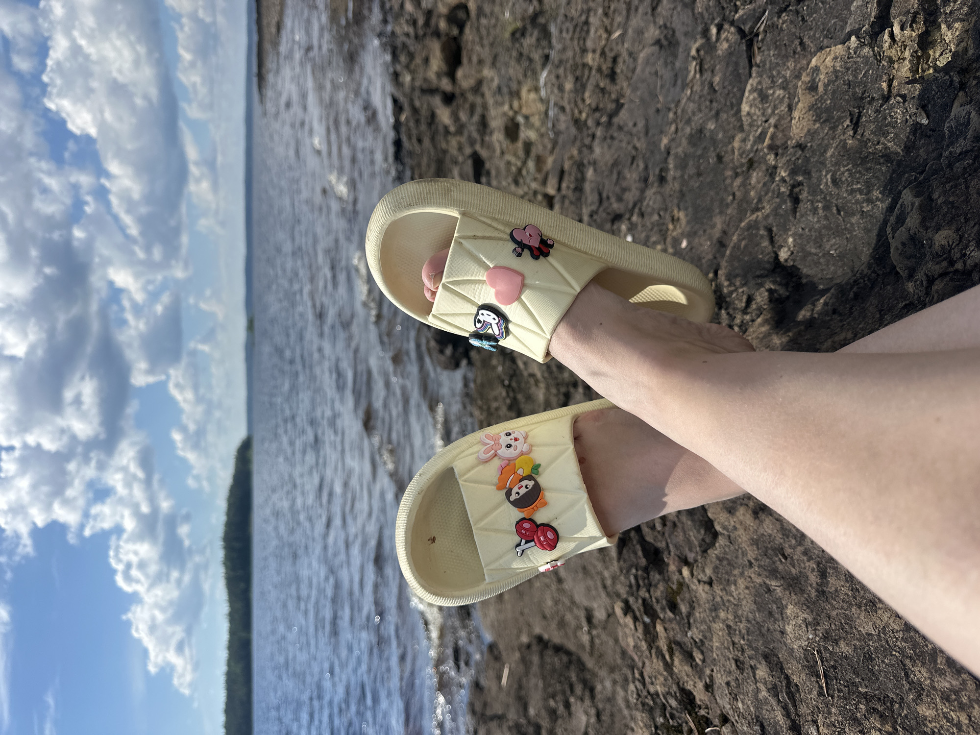 A person's legs are crossed, with yellow slides adorned with decorative charms on their feet. The slides are positioned on a rocky shore beside a body of water. In the background, a distant treeline borders the water under a cloudy sky. The location is identified as Soanlakhti, Russia.