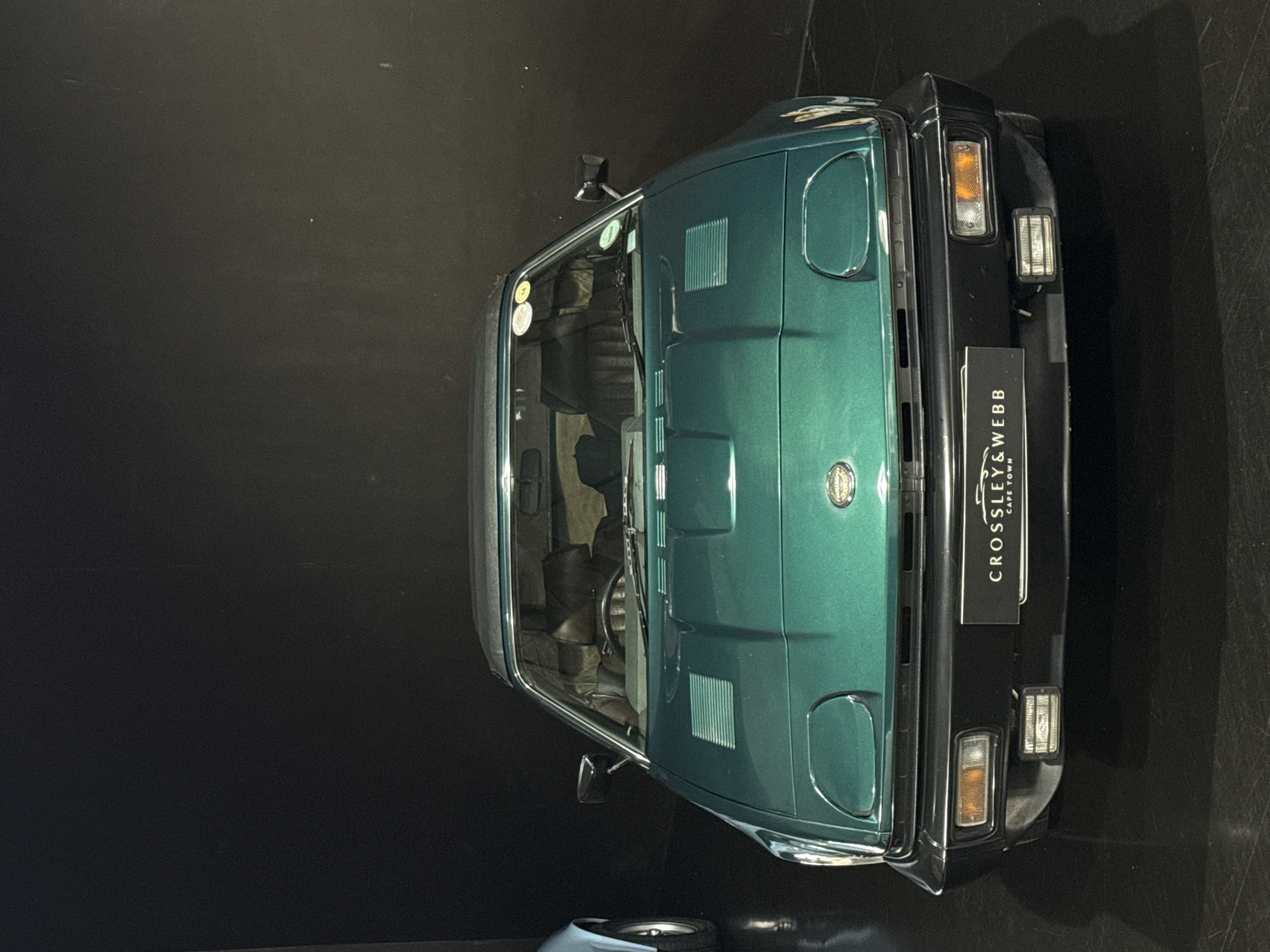 An overhead, slightly angled shot presents a classic metallic green sports car, likely from the 1970s or 1980s, showcased in an indoor setting. The vehicle features a distinctive wedge-shaped design with visible pop-up headlight covers, integrated air vents on the bonnet, and black trim along its edges and bumpers. The car's interior is partially visible through the windscreen, revealing glimpses of the seats and dashboard.
At the front of the car, a prominent black rectangular plate is attached, displaying white text that reads "CROSSLEY & WEBB" at the top and "CAPE TOWN" below it. This detail explicitly places the car's location in Cape Town, South Africa. The background consists of a dark, likely black, and somewhat reflective floor or surface, suggesting a professional display environment such as a showroom, museum, or private collection. The lighting is focused, highlighting the car's contours and metallic paint finish. A small, out-of-focus object, possibly a wheel or part of another vehicle, is visible in the bottom left corner of the frame. The scene is devoid of people or activity, emphasizing the car as a stationary exhibit.