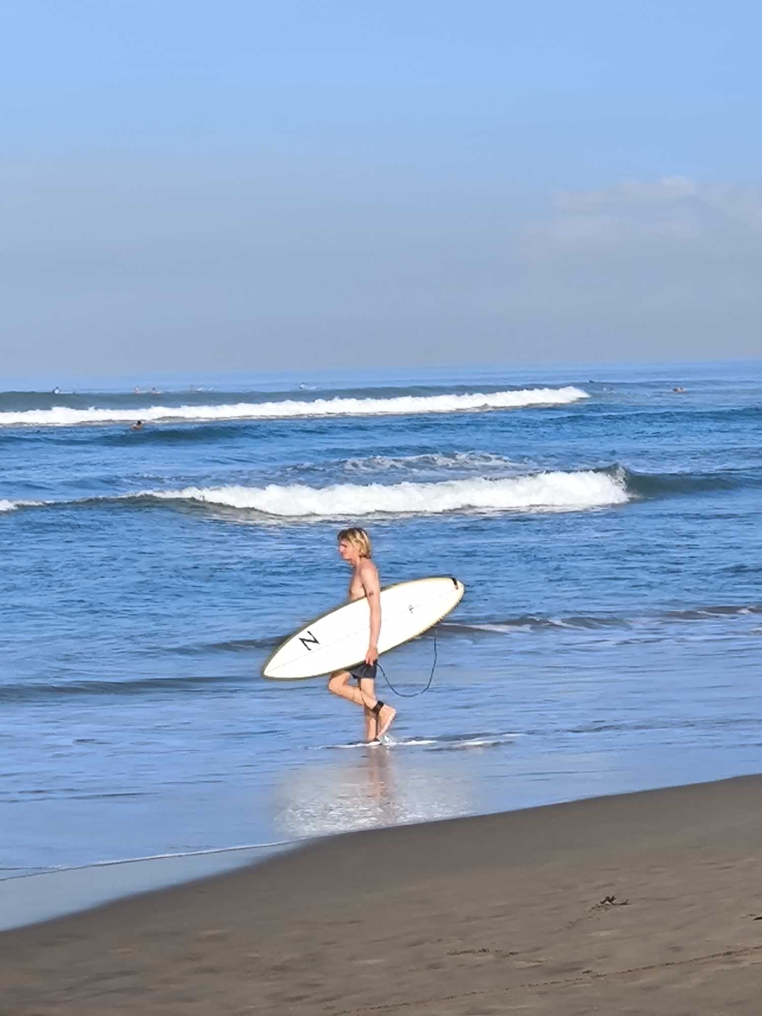 The image depicts a surfer walking in the shallow water along a beach. The location is North Kuta, Indonesia. The surfer, a light-skinned male with blond hair, carries a white surfboard with a black "Z" on it under his arm. He is barefoot and wears dark-colored shorts or swim trunks. The water is blue with small waves breaking in the background. The beach has dark sand. In the distance, other surfers can be seen on the water. The sky is blue with a slight haze.