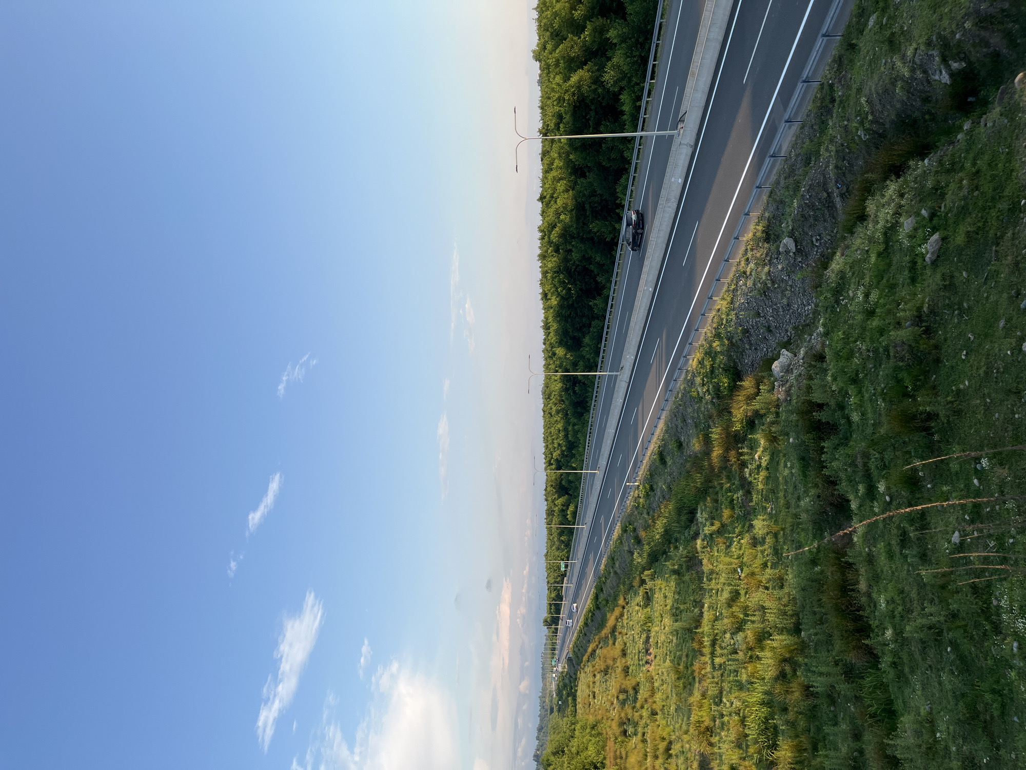 A wide, multi-lane highway stretches into the distance under a clear blue sky with scattered wispy clouds. The highway is bordered by a dense forest of green trees on the right and a grassy, overgrown embankment on the left. Several vehicles are visible on the highway, with one dark SUV in the foreground on the right lane, traveling away from the viewer. Lampposts line the side of the highway, and the grassy area on the left is illuminated by the warm glow of what appears to be late afternoon or early evening sun. The scene is tranquil and suggests a rural or semi-rural setting outside of a city.