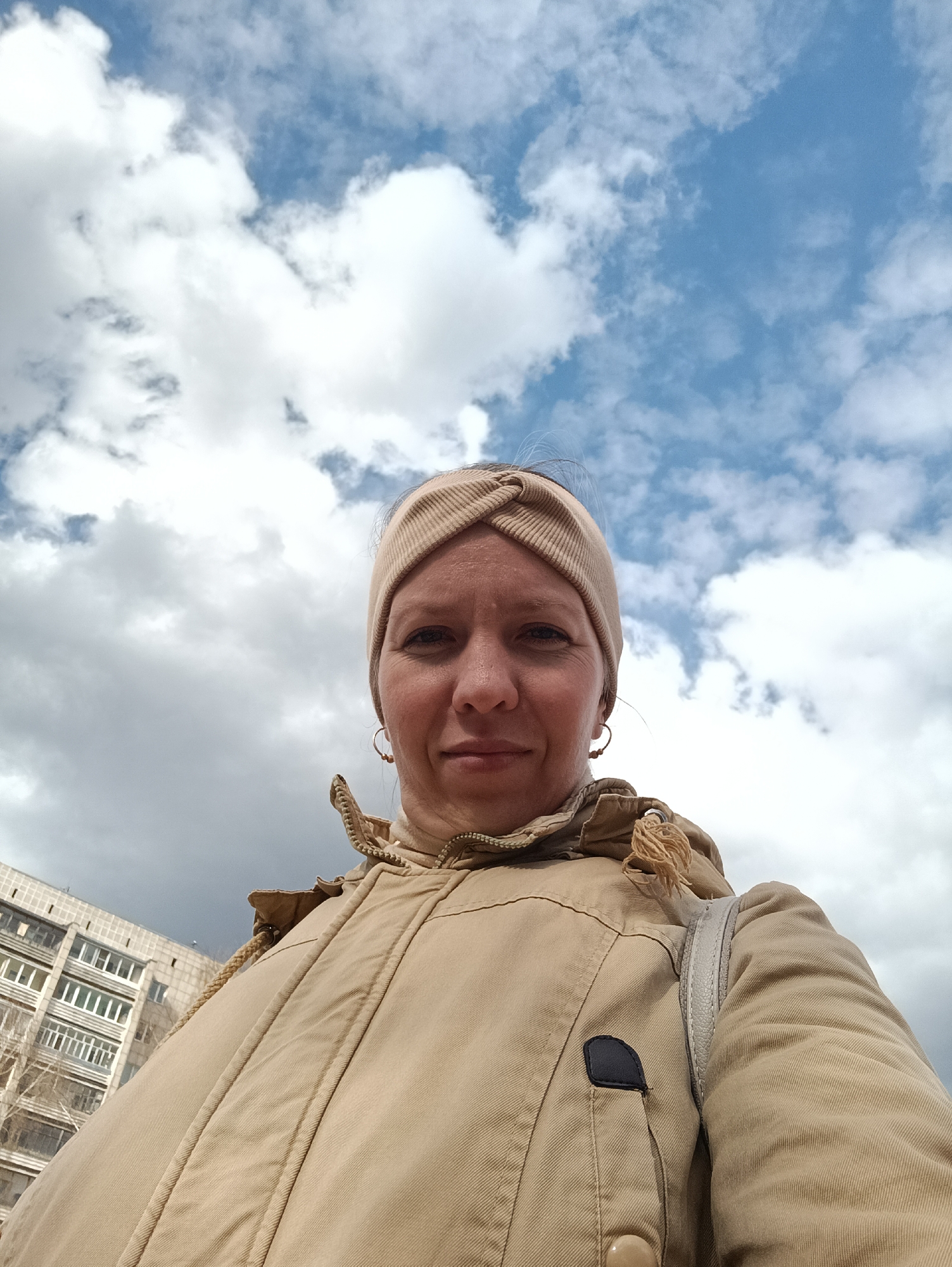 A low-angle outdoor selfie captures a woman in Chaikovsky, Russia. The woman, appearing to be in her 30s or 40s, is centered in the frame, looking directly at the camera with a slight, calm smile. She wears a light beige or tan jacket with a visible zipper and a matching knotted headband. Small hoop earrings are visible on her ears. A light-colored bag strap can be seen over her right shoulder, and a small black patch is on her jacket near the zipper.
The background is dominated by a dynamic sky, featuring both patches of bright blue and large, fluffy white cumulus clouds, along with some darker, more overcast areas. This suggests a mixed weather day. In the lower-left corner, a portion of a multi-story residential building with numerous windows is visible, indicating an urban or suburban setting. The photograph was taken during daytime. No visible text is present in the image.