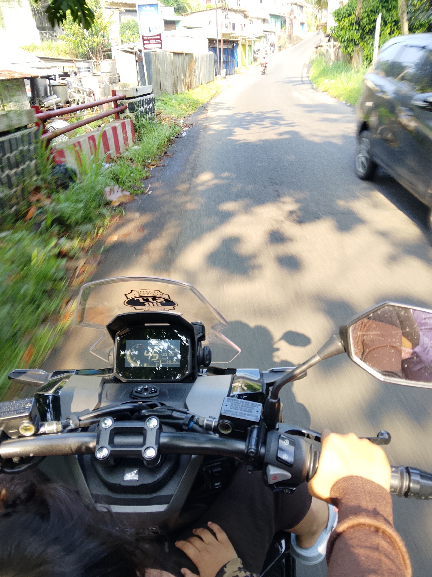 This is a first-person perspective, eye-level shot from a moving motorcycle on a paved road in Cijeruk, Indonesia. The rider's hands are visible on the handlebars. The dashboard display shows the time as 06:00. The road curves to the right, and on the left are buildings and a red-railed barrier. On the right, a black car is moving away from the camera. Trees and foliage line the sides of the road, and shadows from the trees are cast on the road surface. The environment suggests an urban or suburban setting with moderate development.