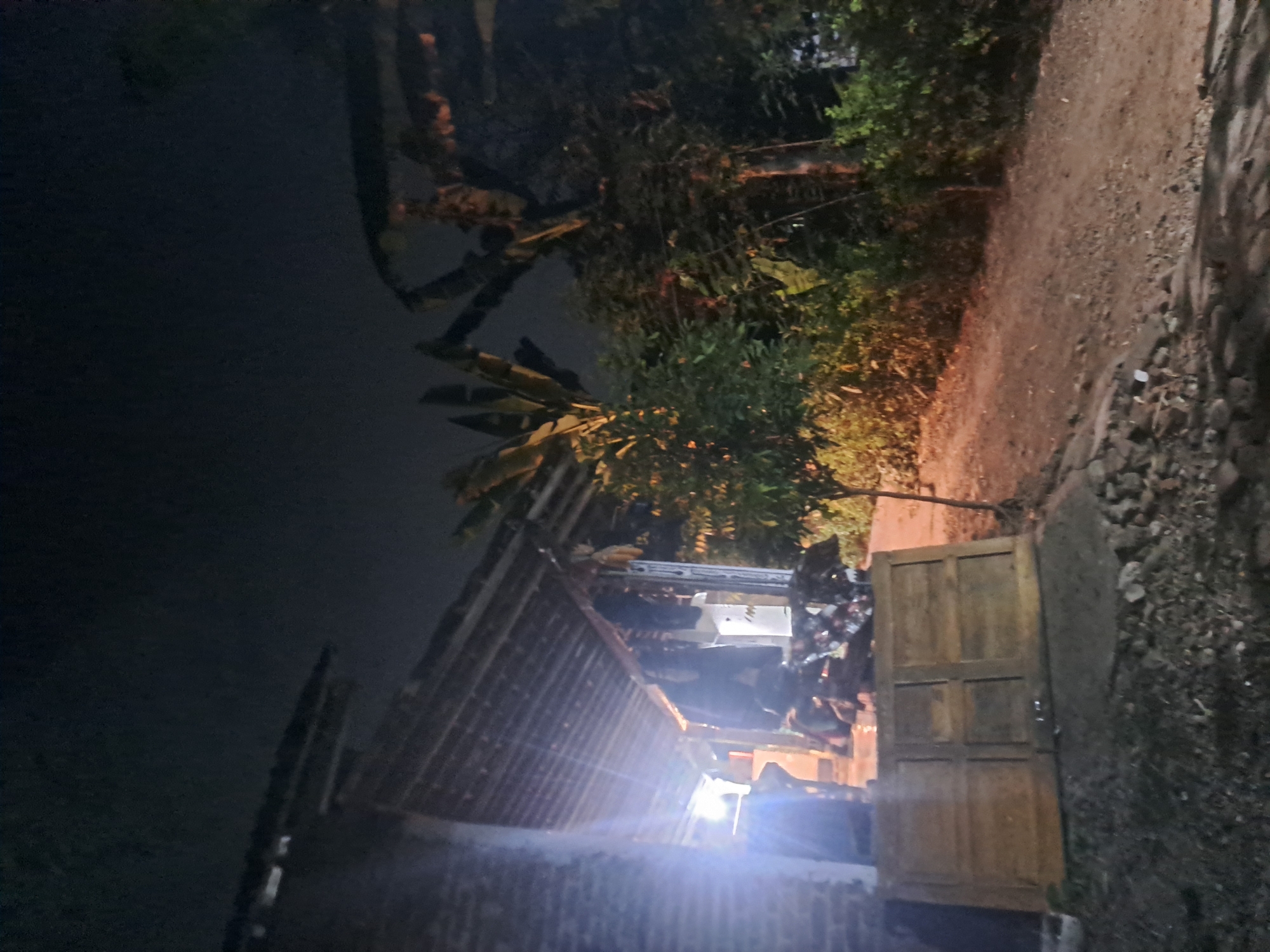 An outdoor night scene is depicted, likely in the city of Sumberejo, Indonesia. On the left, a brick structure with a corrugated roof extends into the frame, with a bright light source illuminating the area beneath the eaves, casting strong glare. Several indistinct human figures are present under the roof. Scattered debris and what appear to be wooden planks are visible in front of the structure. To the right, a dirt path leads away from the building, flanked by dense foliage and trees. The sky is dark and cloudy.