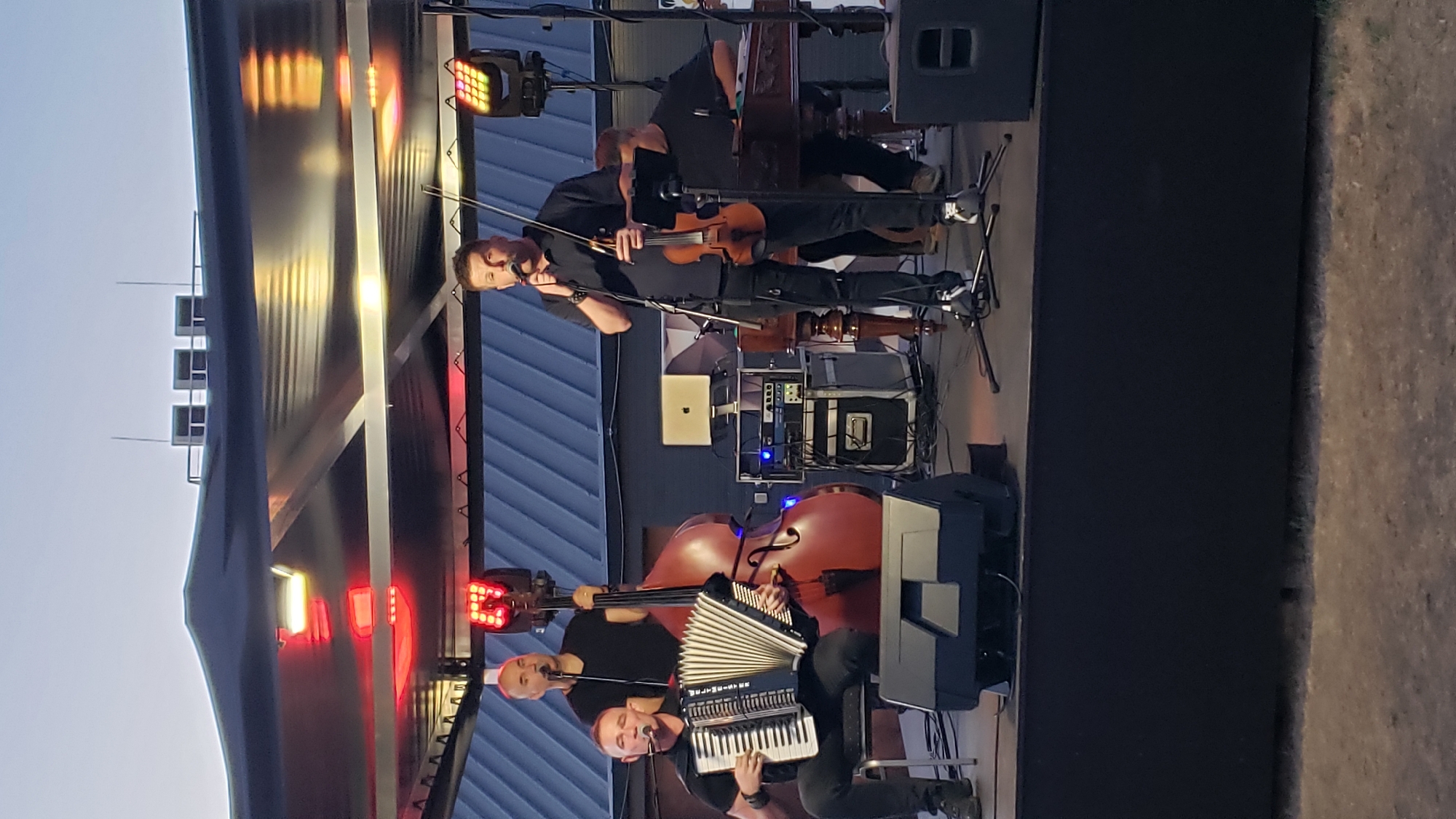 A band is performing on an outdoor stage under a dark canopy. In the foreground, a man sits playing an accordion. To his right, a man stands singing into a microphone and playing a violin. Behind the accordion player, a man stands playing a double bass. Further back, another man is seated, possibly playing a keyboard instrument. The stage is set up with various audio equipment, including speakers, amplifiers, and a laptop. The lighting appears to be a mix of stage lights and ambient evening light. The background shows a blue corrugated metal structure.