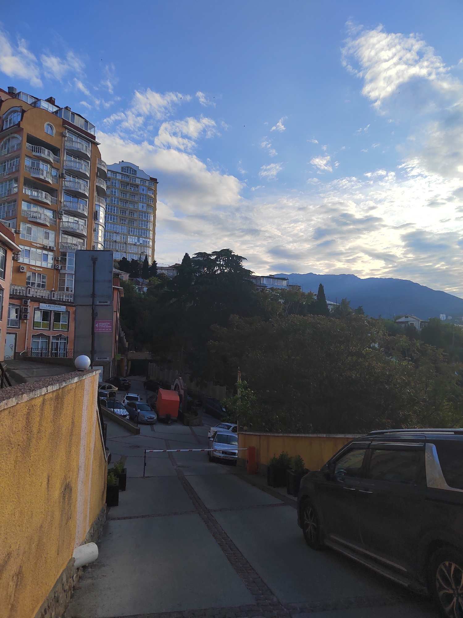 The image shows a street scene in an urban location. The perspective is from an elevated position, looking down a street with a slight slope. On the left and right sides of the street are yellow walls. Several cars are parked along the street. In the background, there are tall buildings with balconies. Behind the buildings, there is a mountain range visible under a partly cloudy sky. The overall lighting suggests it is daytime, possibly late afternoon.