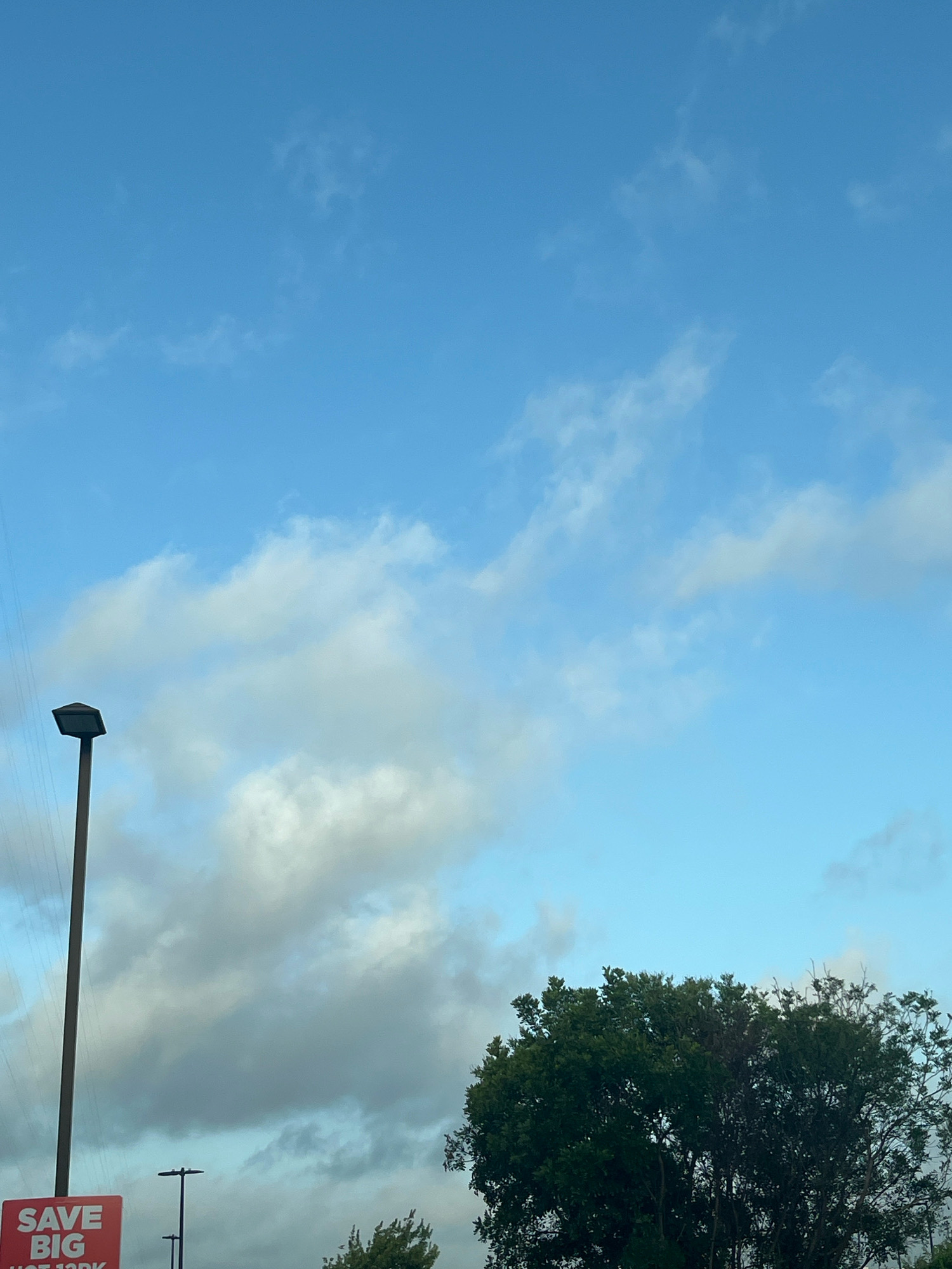 A clear blue sky with scattered white clouds is visible in the upper portion of the image. A tall, dark pole with a light fixture at the top is positioned on the left side, extending vertically. Below the light pole, a red sign with white text that reads "SAVE BIG" is partially visible at the bottom left corner. A dense green tree occupies the lower right quadrant of the image. The scene appears to be outdoors in an urban environment, potentially San Antonio, United States of America, given the provided context. No people or specific actions are depicted.