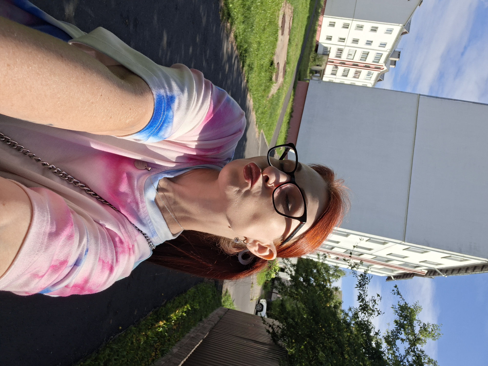 A selfie taken from a low angle shows a young woman with long, auburn hair pulled back in a ponytail, wearing glasses and a tie-dye t-shirt in shades of pink, blue, and white. She is standing outside on a paved walkway, with green grass and apartment buildings in the background. The apartment buildings are white with pink accents and appear to be located in a residential area of Myadzyel, Belarus. The sky is blue with scattered white clouds. The woman is pouting her lips, and the photo is taken in daylight.
