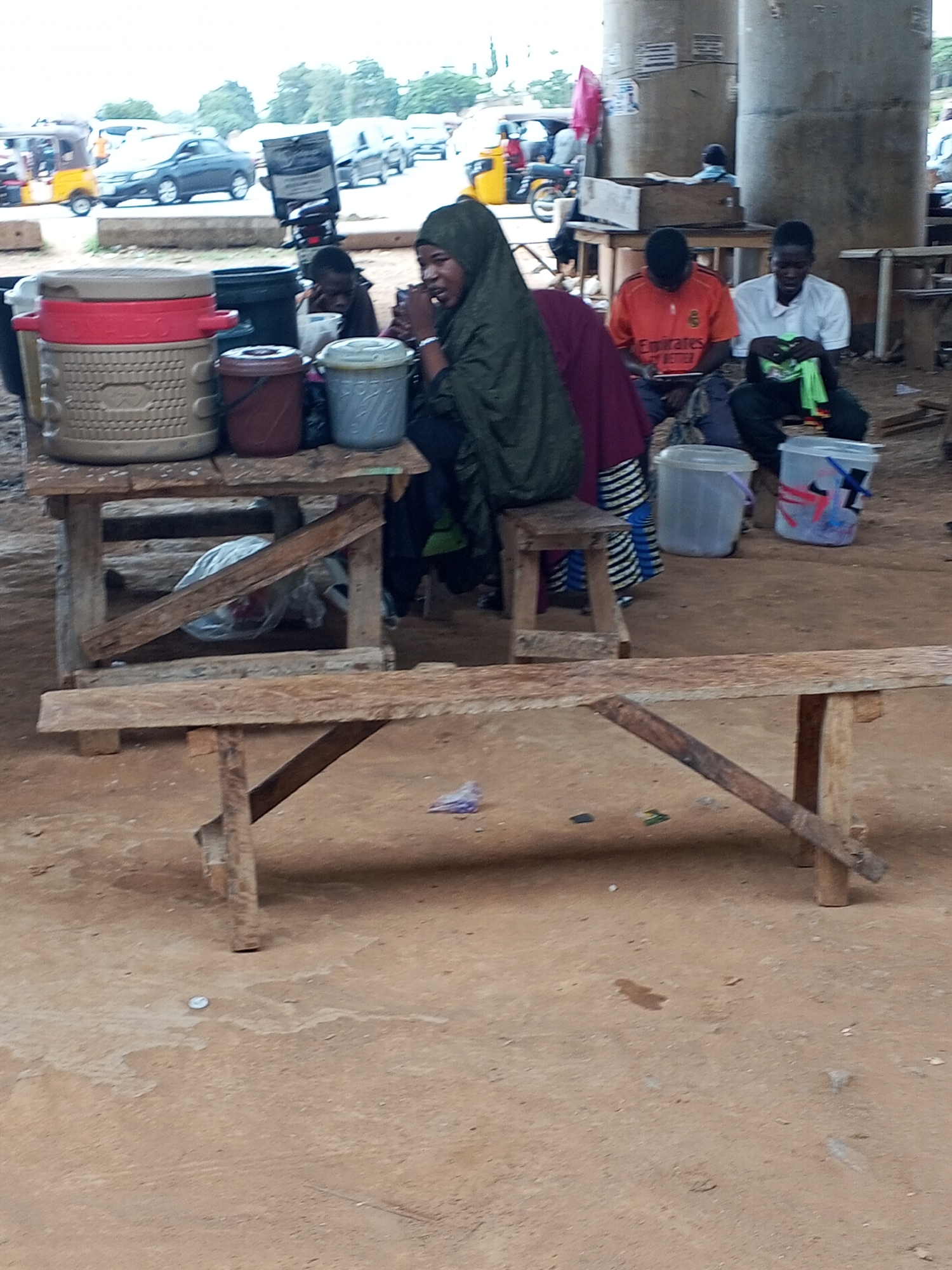 The image shows a scene under a bridge in Galadimawa Village, Nigeria. Four people are visible, two are seated at a table with several buckets and containers on it. A woman in a green hijab sits on a stool. To her right, two men are seated; one wears an orange shirt and looks at a phone or tablet, while the other wears a white shirt and appears to be sewing or repairing something. There is a wooden bench in the foreground and a few vehicles and motorcycles visible in the background on the other side of the underpass. The ground is dusty and has some trash scattered on it. The scene appears to be an informal gathering or a makeshift market.