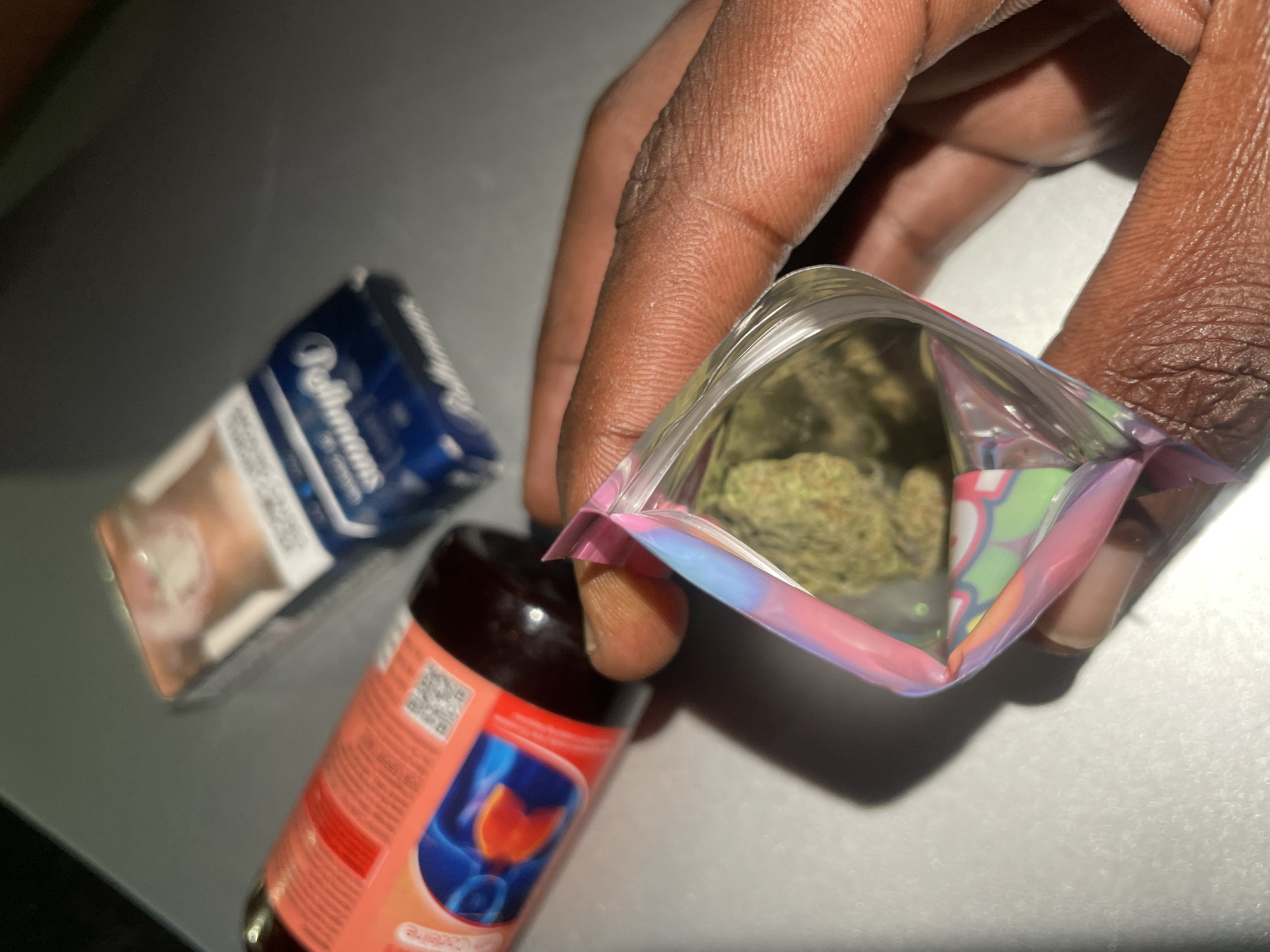 The image shows a dark-skinned hand holding open a small, resealable plastic bag containing what appears to be cannabis buds. A pack of cigarettes is visible in the background, along with a bottle of cough syrup. The setting is a dark surface, possibly a table. There is no indication of the city of Abuja, Nigeria, within the visual data provided.