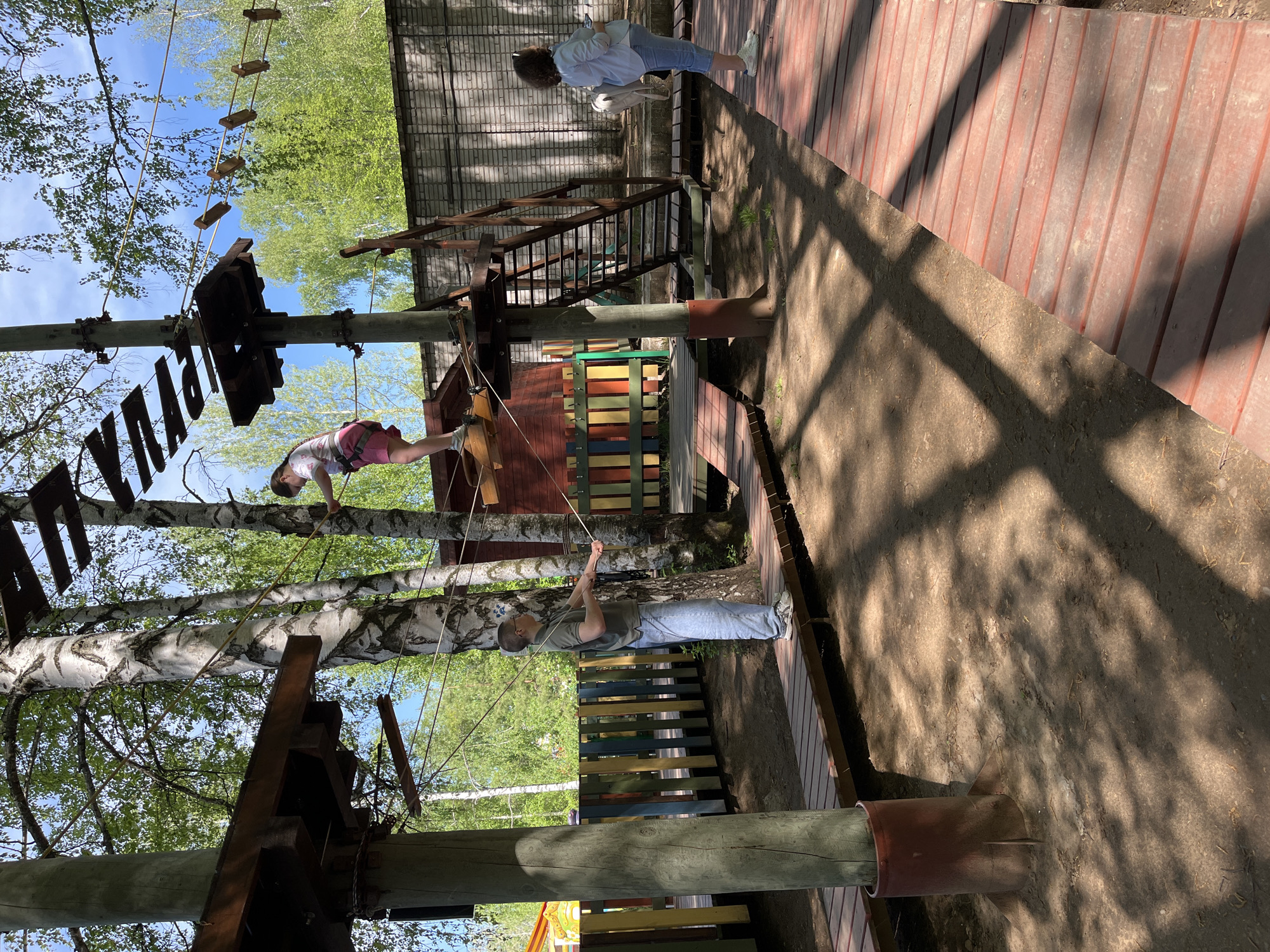 The image captures a scene at a rope park in Chaikovsky, Russia. A girl is seen navigating an obstacle course high in the trees, while a man stands below, possibly assisting or supervising her. Another person is visible on a wooden walkway, further away from the main action. The park features wooden platforms, ropes, and other climbing elements attached to trees, creating an adventurous environment. The lighting suggests a sunny day, with dappled shadows cast on the ground and structures. The background includes lush greenery and a partial view of a building.