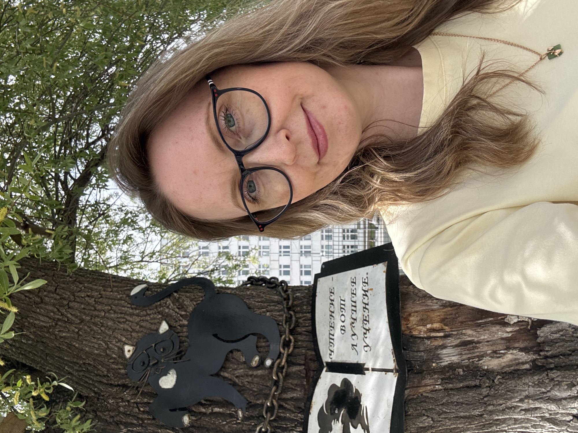 A young woman wearing glasses and a light yellow shirt poses for a photograph outdoors. She is standing near a tree with a metal cat wearing glasses attached to the bark. Next to the cat is a sign in Russian. The text on the sign reads, "Чтение вот лучшее учение", which translates to "Reading is the best learning." The location is Chaikovsky, Russia. The backdrop includes a tree with green leaves and a multi-story building in the distance. The lighting suggests it is daytime.