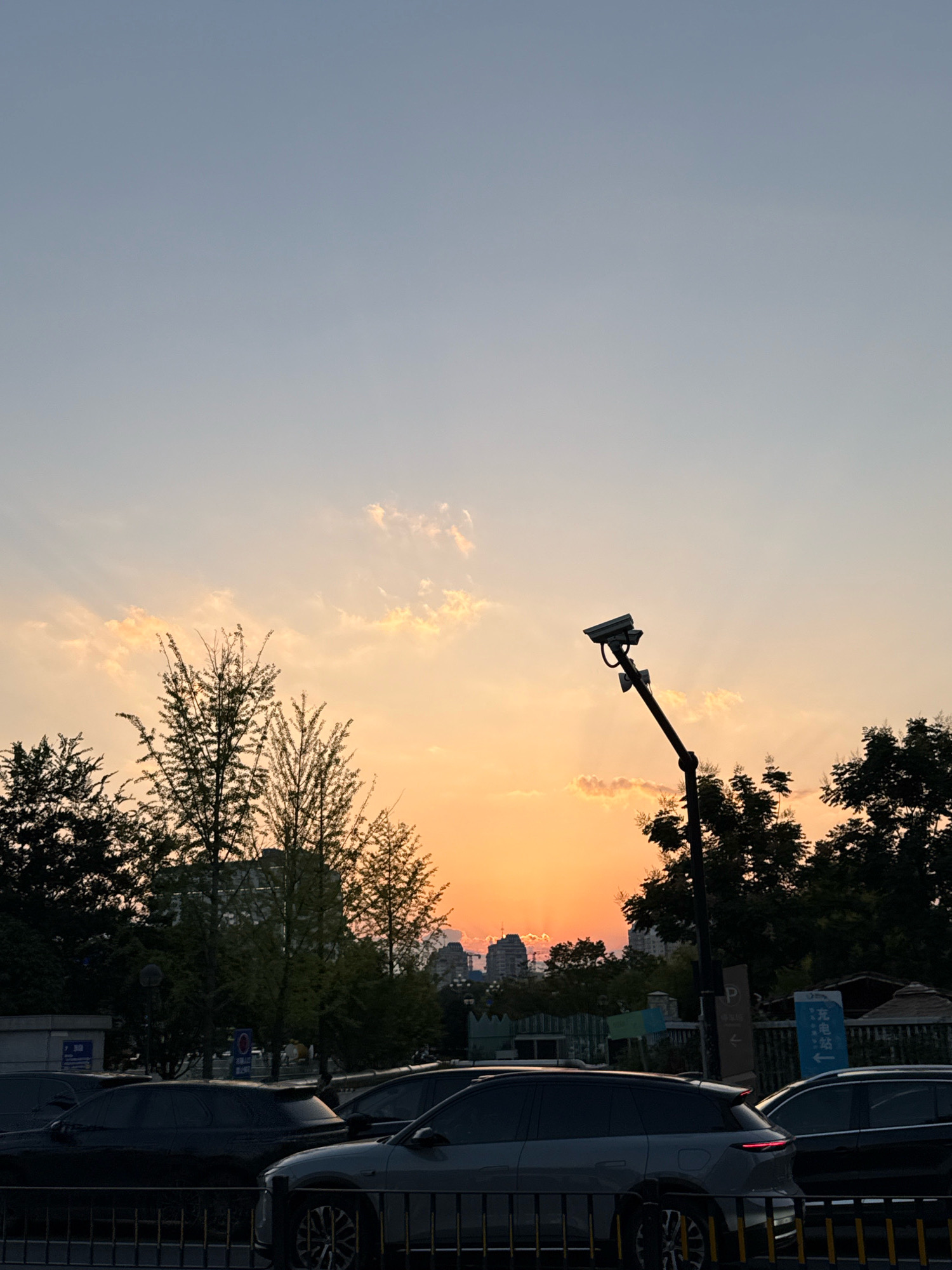 The image depicts a street scene in Yiwu, China, during sunset. The sky is a gradient of pale blue to warm orange, with scattered clouds tinged by the setting sun. Silhouetted trees frame the lower portion of the sky, their branches appearing delicate against the vibrant sky.
In the mid-ground, a street lamp with two surveillance cameras is prominently visible, angled towards the left side of the frame. Beyond the trees, the rooftops and some buildings of the city are visible.
In the foreground, several cars are parked or moving along the street, indicated by their headlights and taillights. A dark colored SUV in the center foreground has its brake lights illuminated. A metallic fence runs along the bottom of the image, separating the street from a pedestrian area or sidewalk. The overall atmosphere suggests an urban environment at dusk.