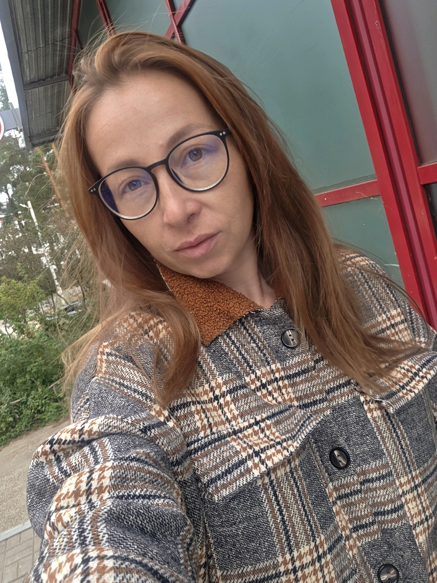 A woman with auburn hair and glasses is taking a selfie outdoors. She is wearing a plaid jacket with a textured collar. Behind her, a building with green siding and red trim is visible, along with some trees and foliage. The location appears to be the city of Chaikovsky, Russia.