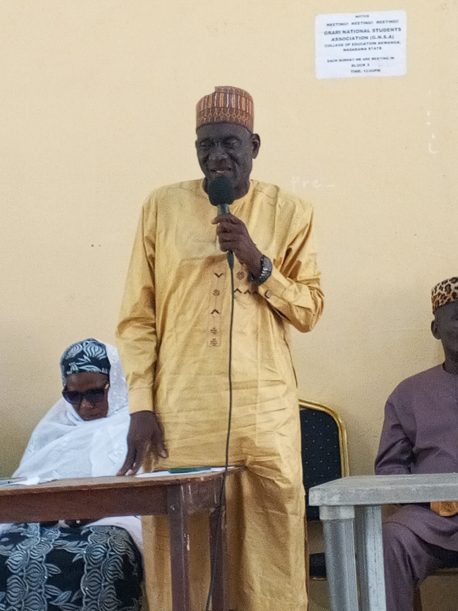 The image shows an indoor scene. A man in a gold tunic and patterned hat speaks into a microphone. He is positioned in front of a light-colored wall with a posted notice: "MEETINGT MIKETING MARTINOS, GRARI NATIONAL STUDENTS ASSOCIATION (G.N.S.A), COLLEGE OF EDUCATION AKWANGA, NASARAWA STATE...BLOCK 3." Seated at a table to the man's left is a woman wearing sunglasses and a white head covering. To the man's right, another man in a purple garment and hat sits at a separate table. The location is likely the College of Education in Akwanga, Nigeria, given the text on the notice.